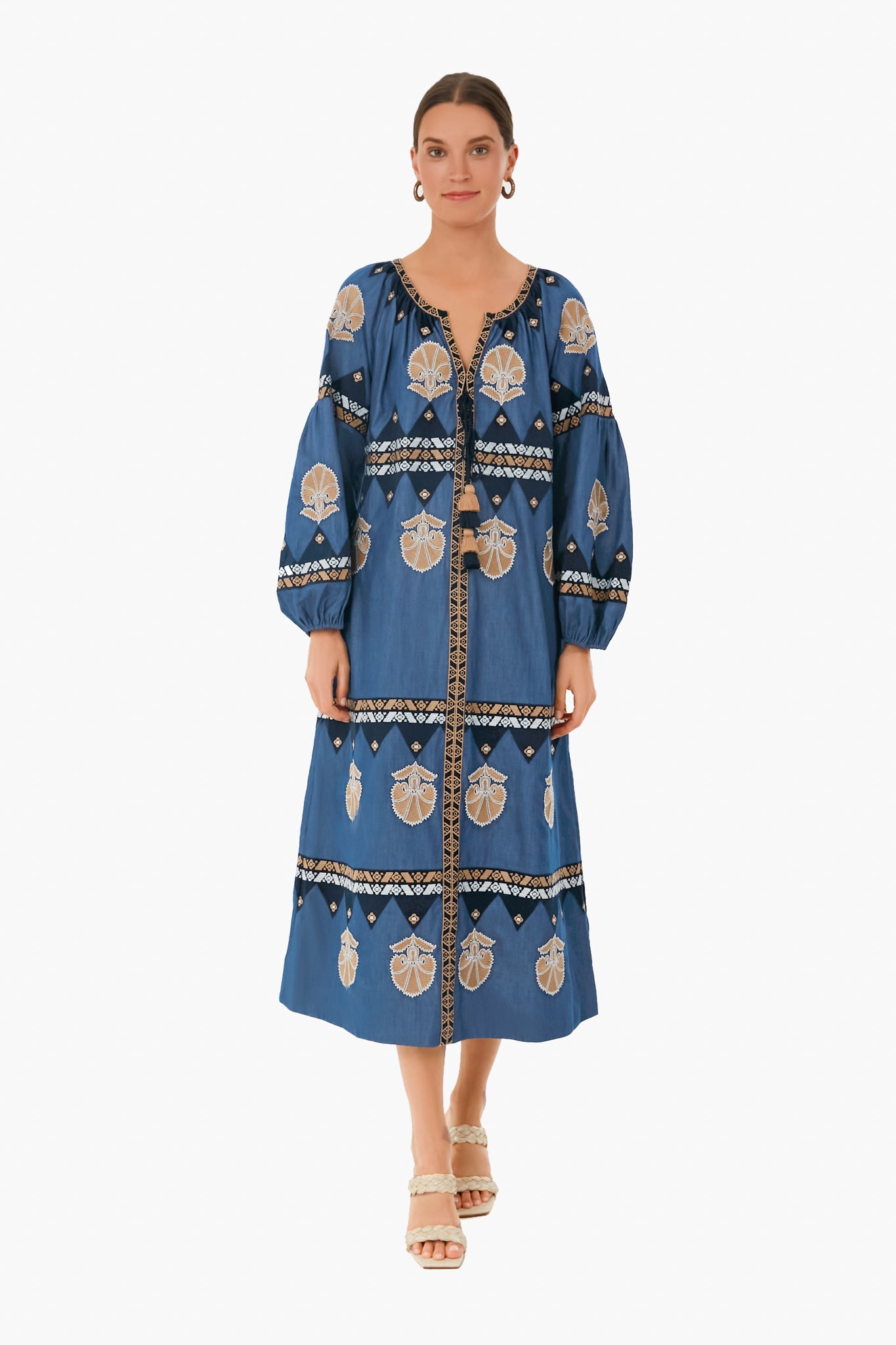 A woman faces forward in a chambray blue Figue long-sleeve embroidered midi dress with beige and black patterns, paired with beige heeled sandals.