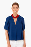 Wearing the XiRENA Tiara Blue Jules Top with a ruffled neckline and bubble sleeves, paired with white pants and a chunky red beaded necklace, she smiles confidently at the camera.