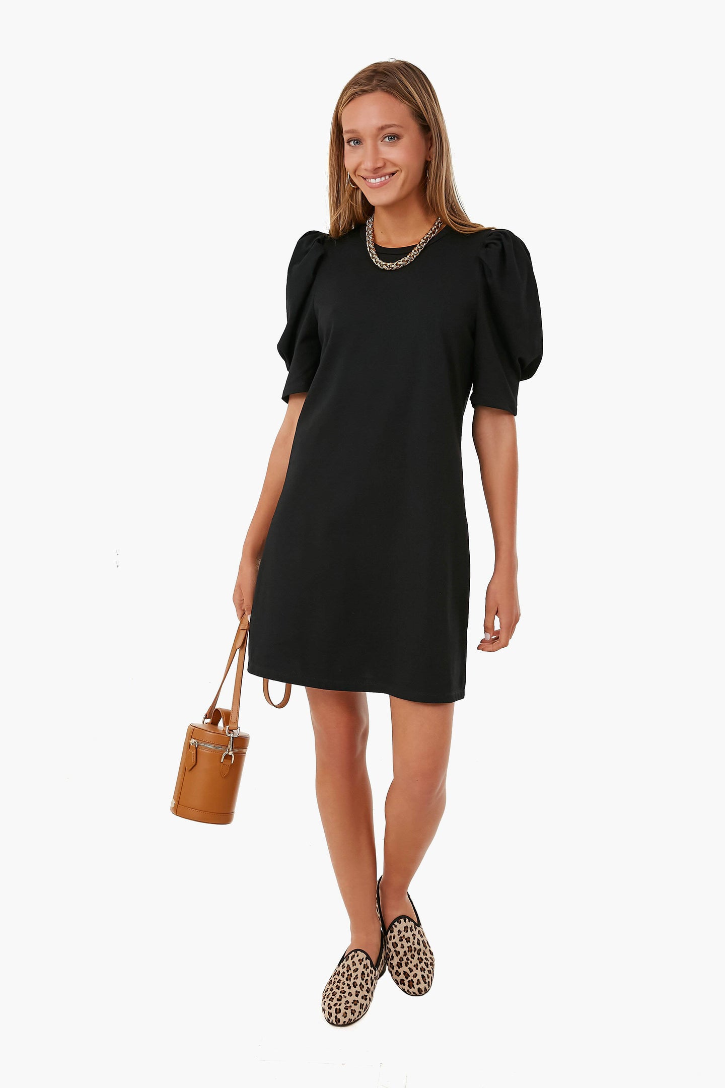 A woman wears a black Pomander Place Stella Dress with puff sleeves, leopard print loafers, a chain necklace, and carries a tan bucket bag.