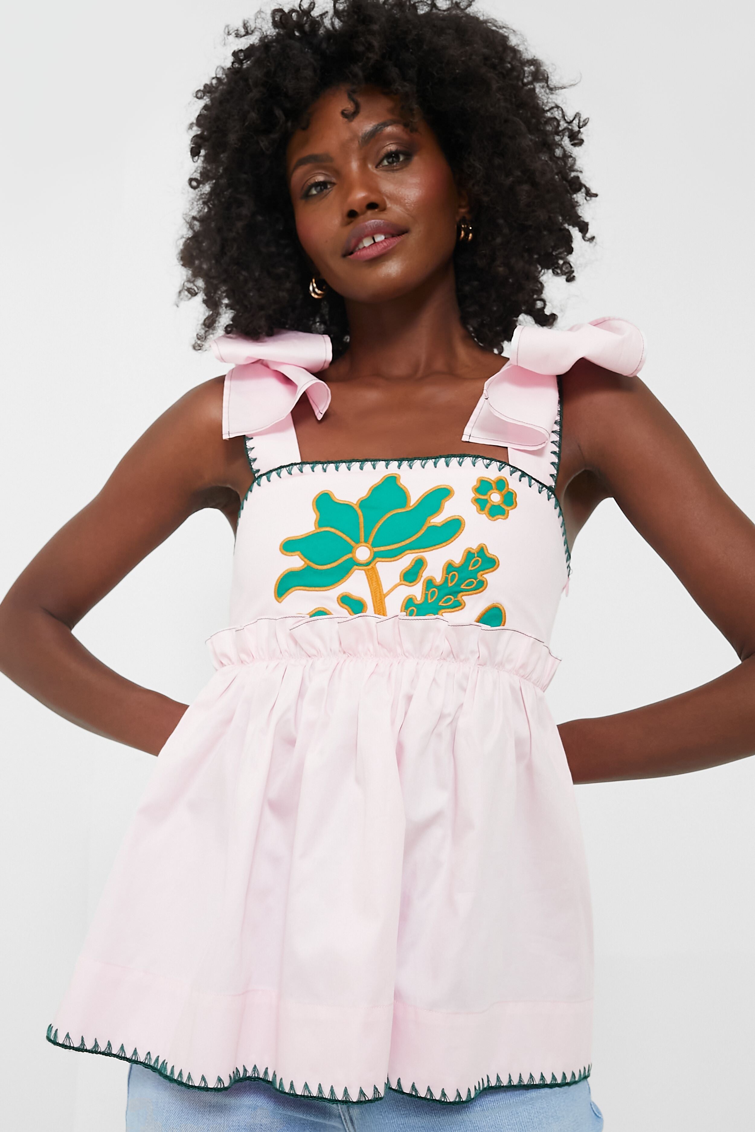 A woman wears the Lug Von Siga Perla Top, a sleeveless pink and green piece featuring shoulder bows and green floral embroidery.