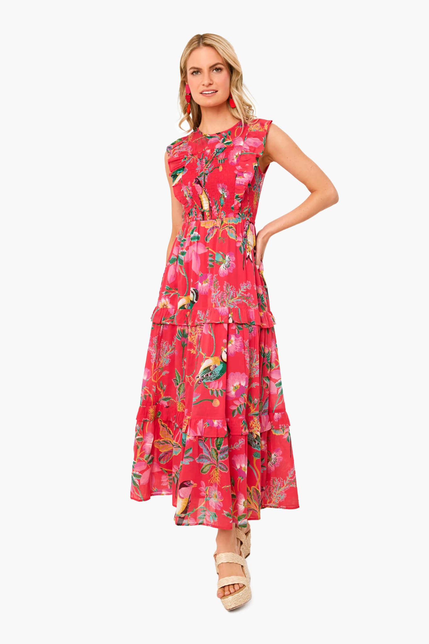 A woman poses in the Banjanan Hedgerow Paradise Pink Iris Dress, a sleeveless ankle-length floral maxi made from organic cotton with ruffle details and exquisite handmade embroidery, paired with wedge sandals.