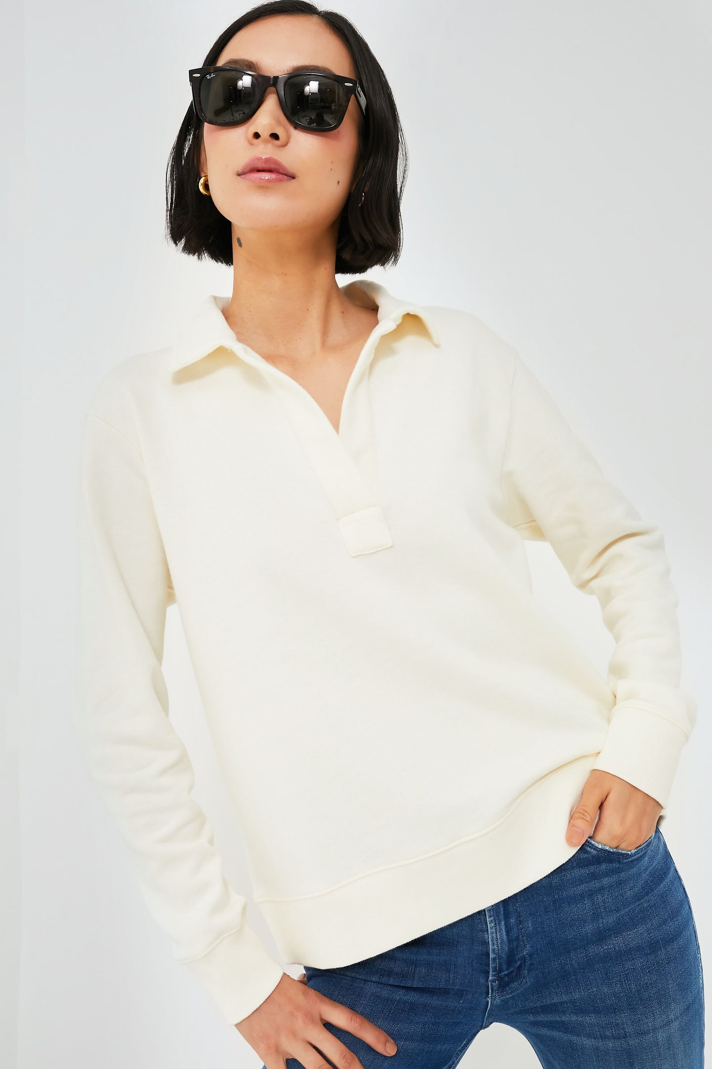 A woman wearing black sunglasses, a cream-colored Vineyard Vines Marshmallow Polo Popover Sweatshirt, and blue jeans stands with one hand in her pocket.
