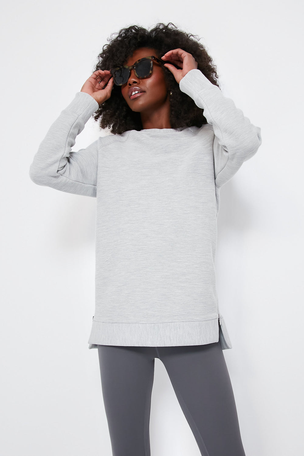 A person in a light gray Varley oversized sweatshirt crafted from ottoman rib fabric and dark gray leggings adjusts their black sunglasses.