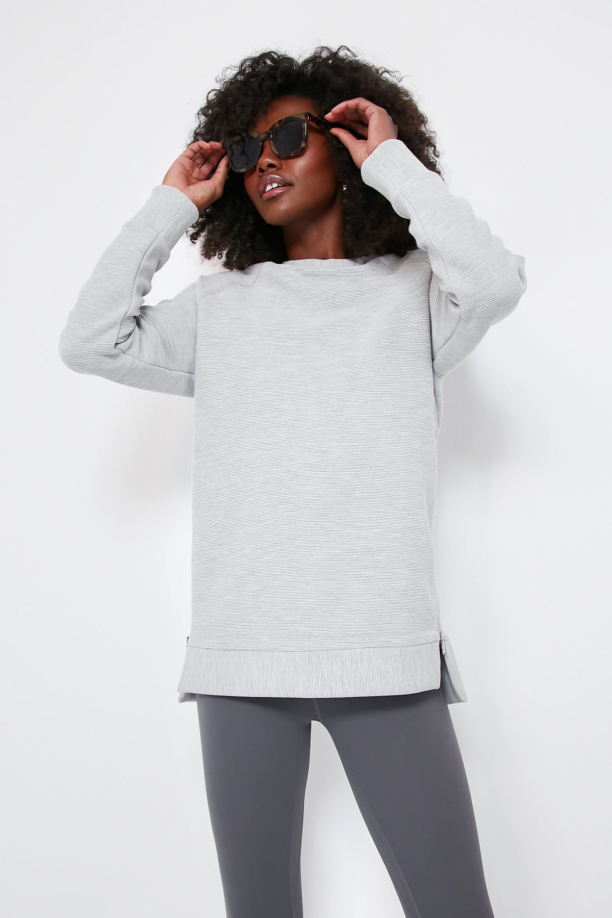 A person in a light gray Varley oversized sweatshirt crafted from ottoman rib fabric and dark gray leggings adjusts their black sunglasses.