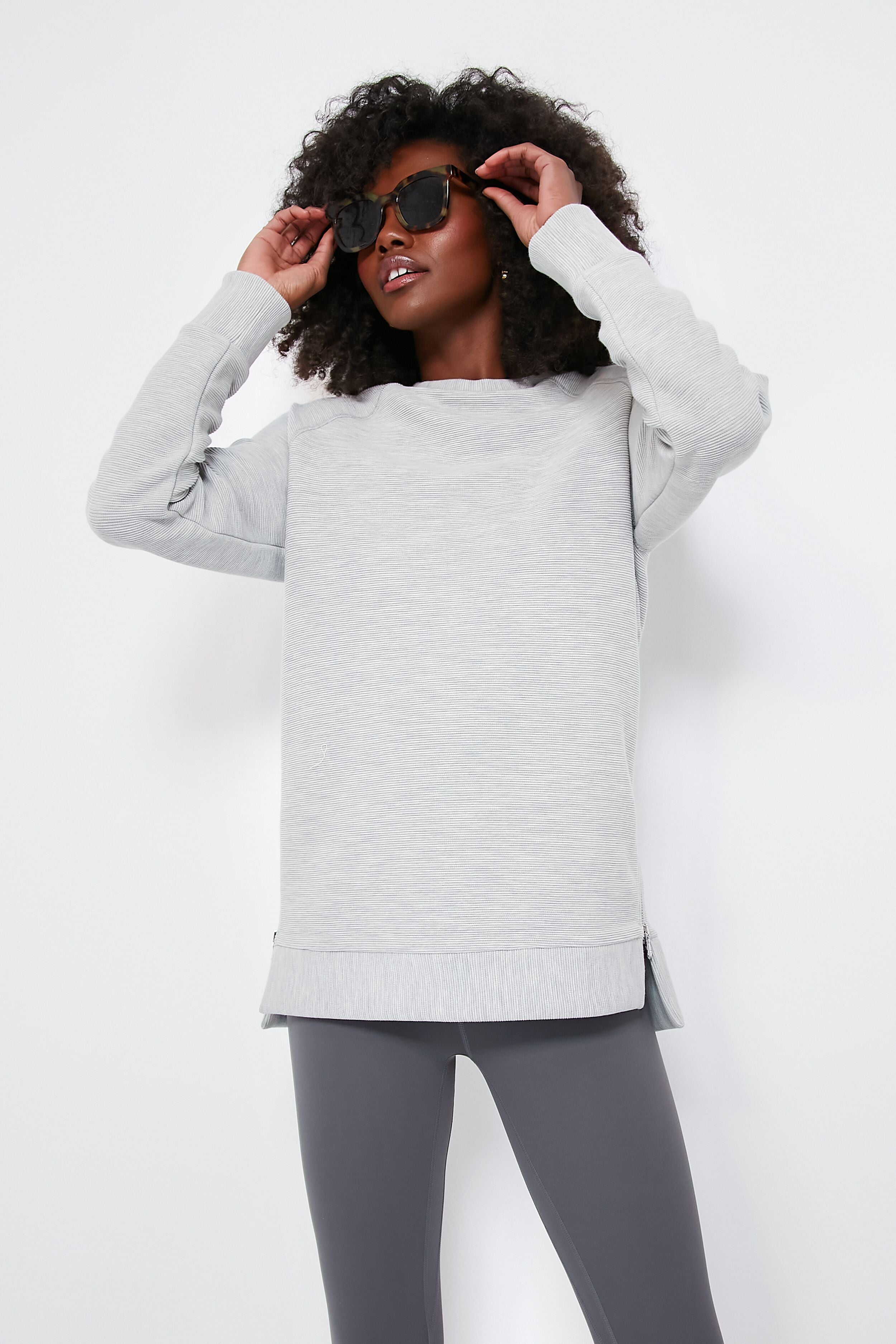 A person in a light gray Varley oversized sweatshirt crafted from ottoman rib fabric and dark gray leggings adjusts their black sunglasses.