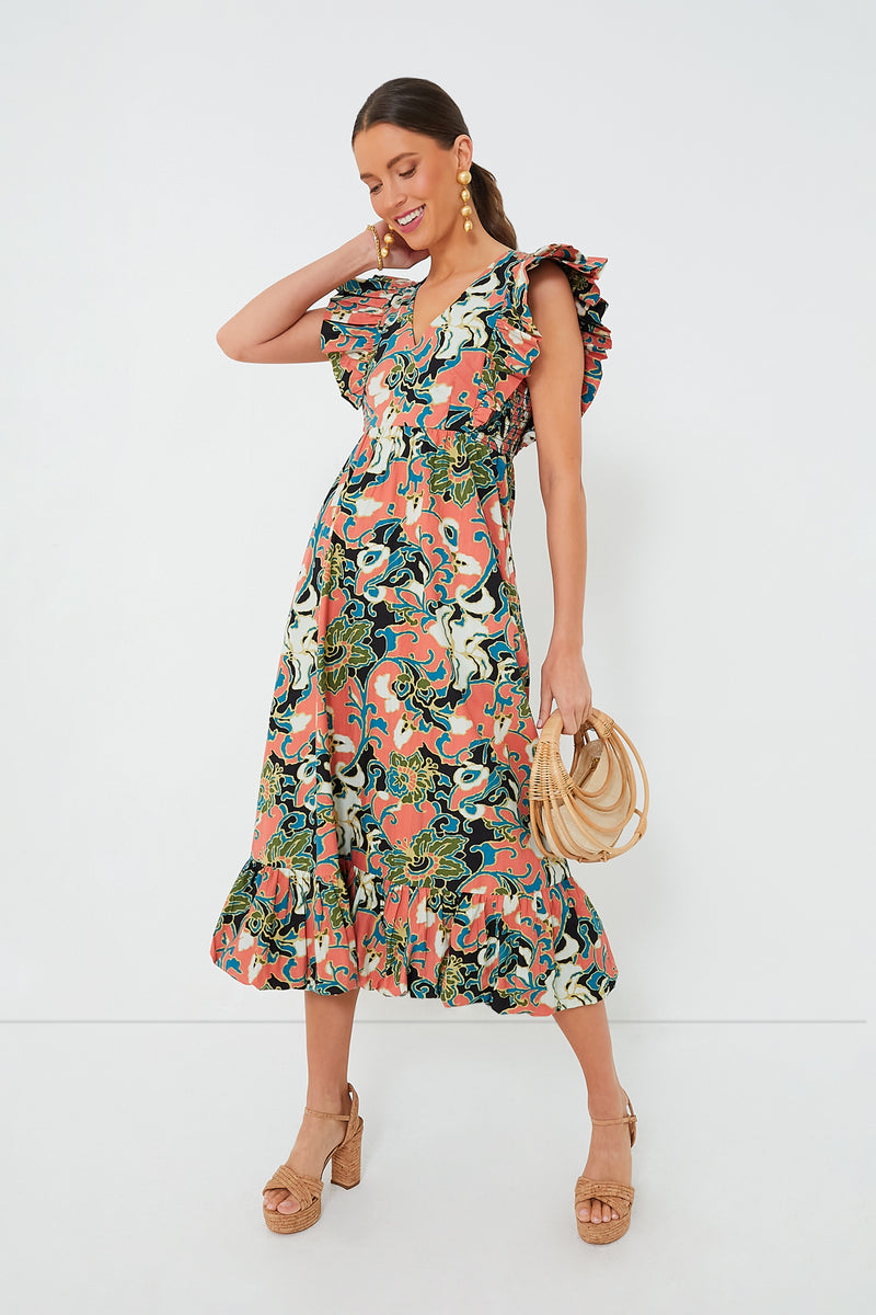 A woman wears Cleobella's Gypsy Bloom Barbara Midi Dress, an ethically handmade piece with colorful patterns and ruffled sleeves. She pairs it with tan platform sandals and carries a round straw bag.
