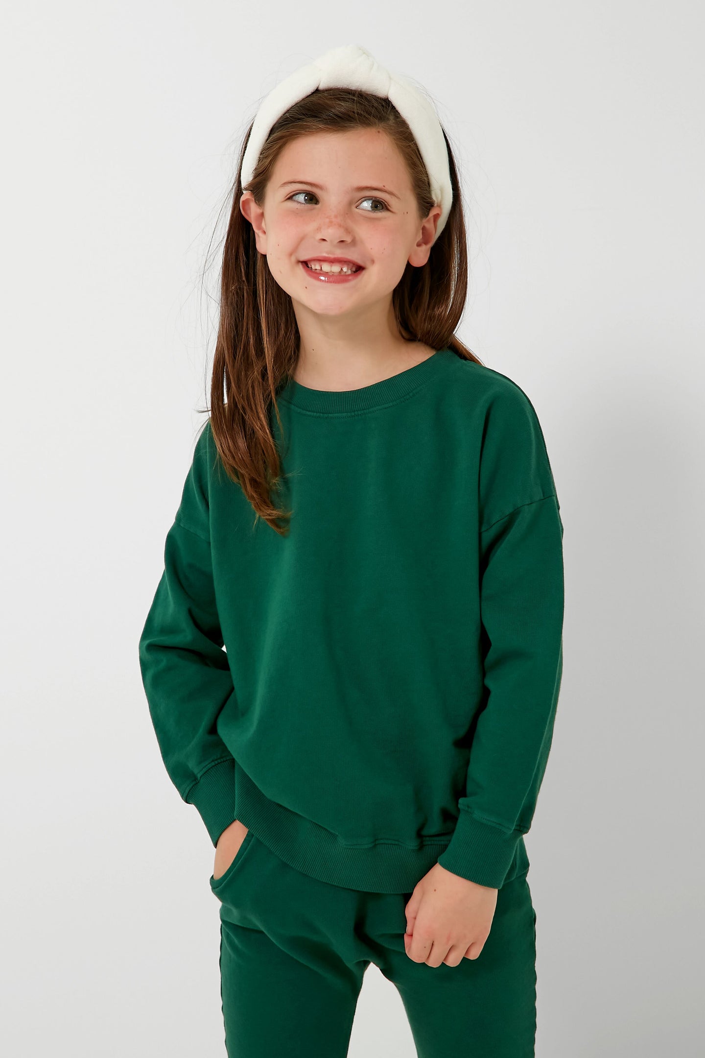 A young girl with long brown hair, a white headband, and a green organic cotton sweatshirt with matching pants smiles and looks to the side. She shines in Les Gamins’ sustainable kids clothing, ethically made in LA.