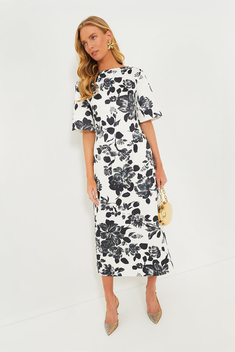 A woman in a white midi Emilia Wickstead dress with black floral print, gold earrings, a small gold purse, and pointed heels exemplifies modern femininity.
