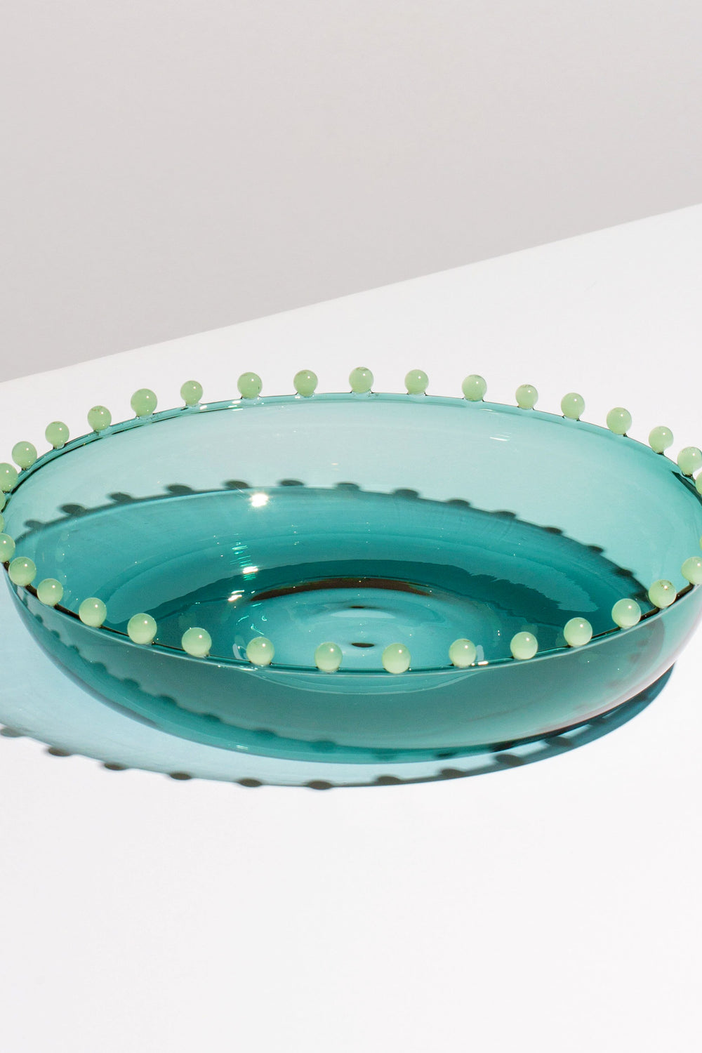 A shallow, translucent blue-green FAZEEK bowl made of hand-blown borosilicate glass with small round beads around the rim rests on a white surface.