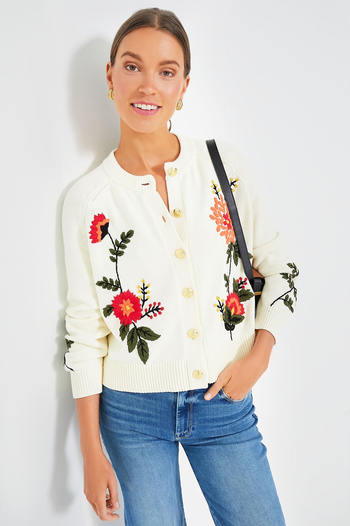 Woman wearing the Tuckernuck Quincy Cardigan in cream with embroidered florals, blue jeans, hoop earrings, and a black shoulder bag.