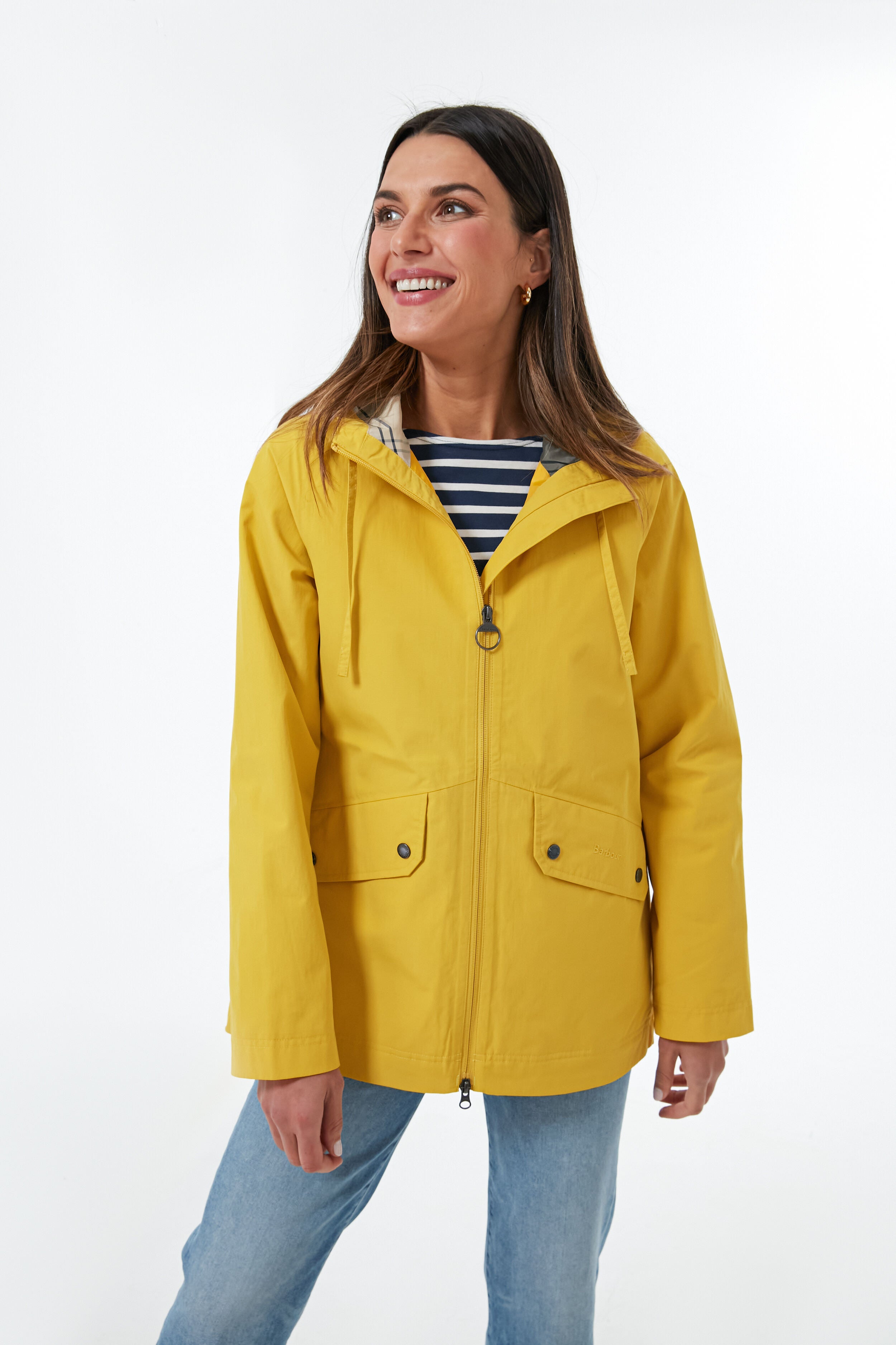 A woman stands confidently in a Barbour yellow waterproof raincoat over a navy and white striped shirt and light blue jeans.