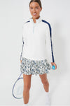 A woman wears a white Tnuck Sport Carver Quarter Zip long-sleeve top and floral pleated skirt, holding a tennis racket and blue ball, showcasing the outfit’s cooling fabric and roomy fit.