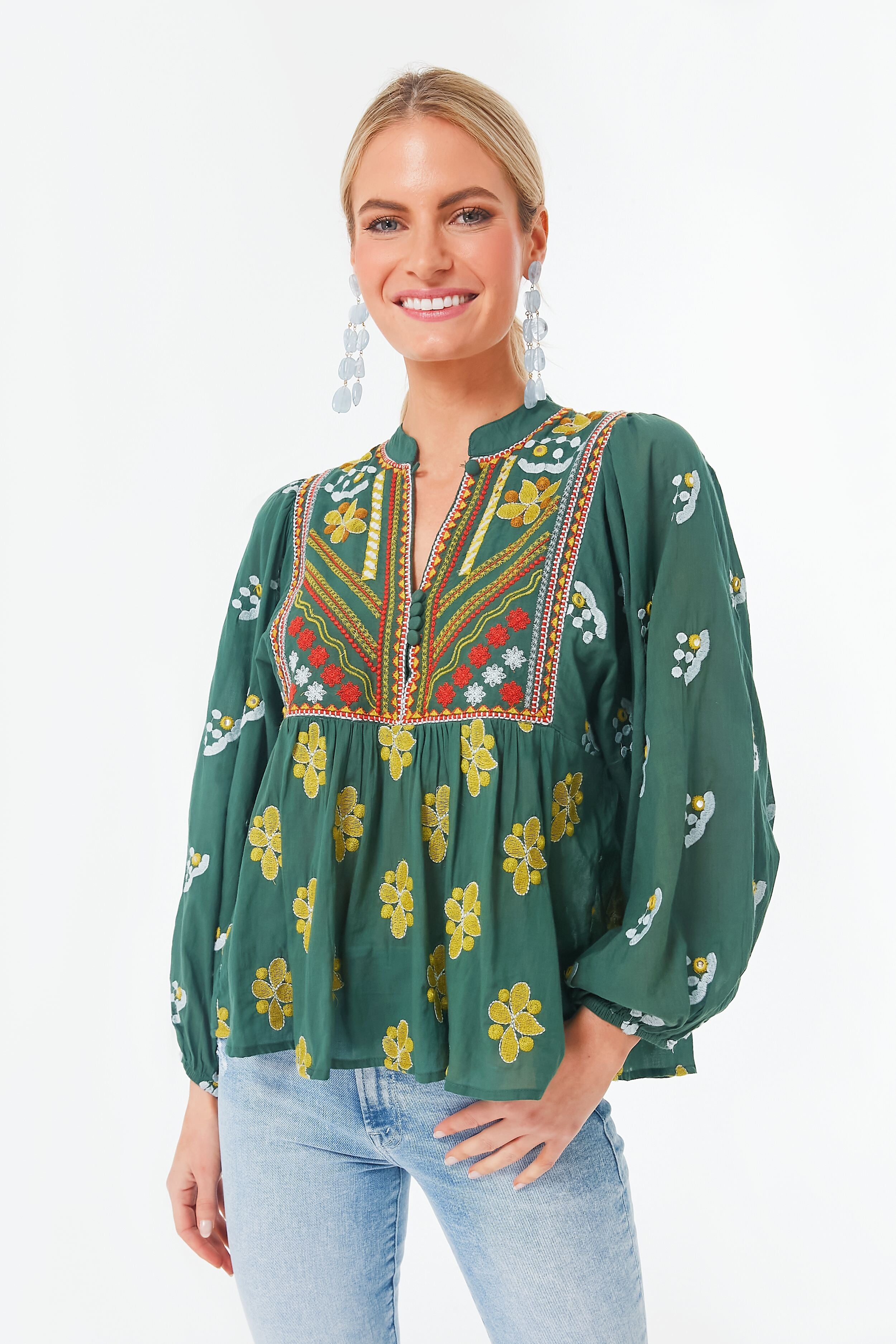 A woman wears an Oliphant green bohemian blouse with floral patterns and colorful embroidery, paired with light blue jeans and drop earrings.