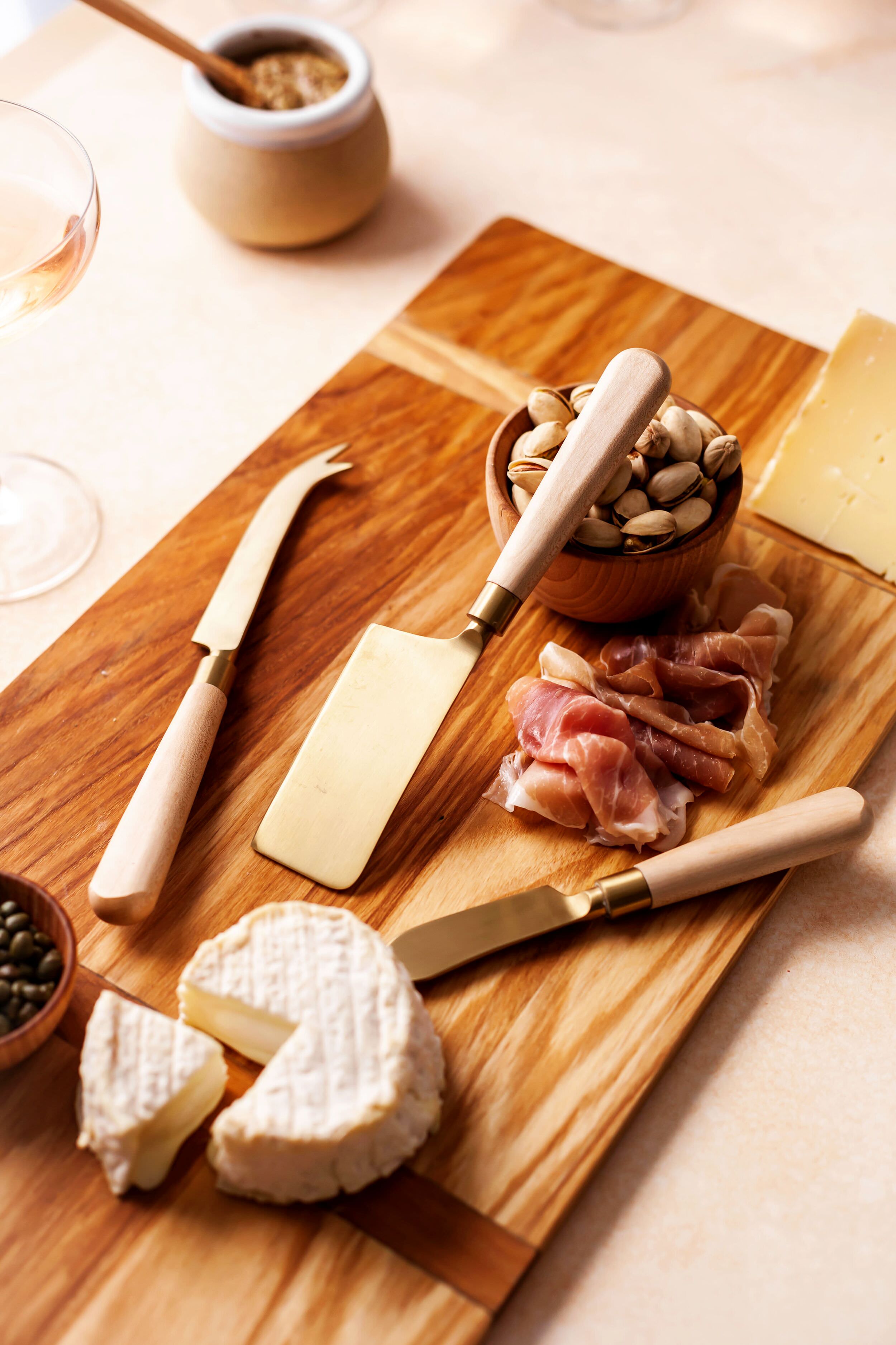 A handcrafted Vermont charcuterie board by Farmhouse Pottery, Inc features cheese knives, a wedge of soft cheese, sliced cured meat, pistachios, capers, and a block of hard cheese.