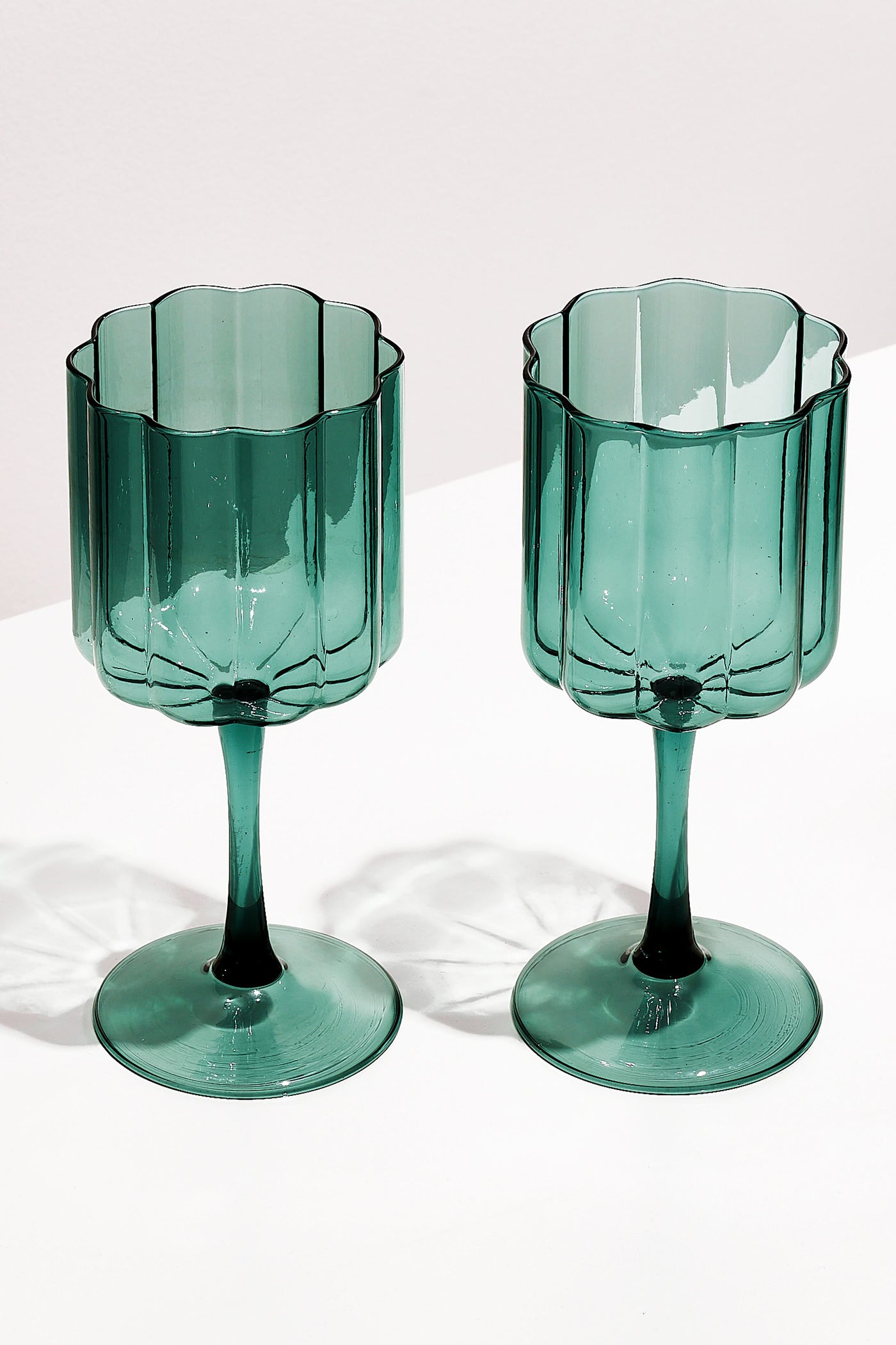 Two FAZEEK Teal Wave Wine Glasses with green-tinted, scalloped edges and textured sides, crafted from hand-blown borosilicate glass, rest on a white surface.
