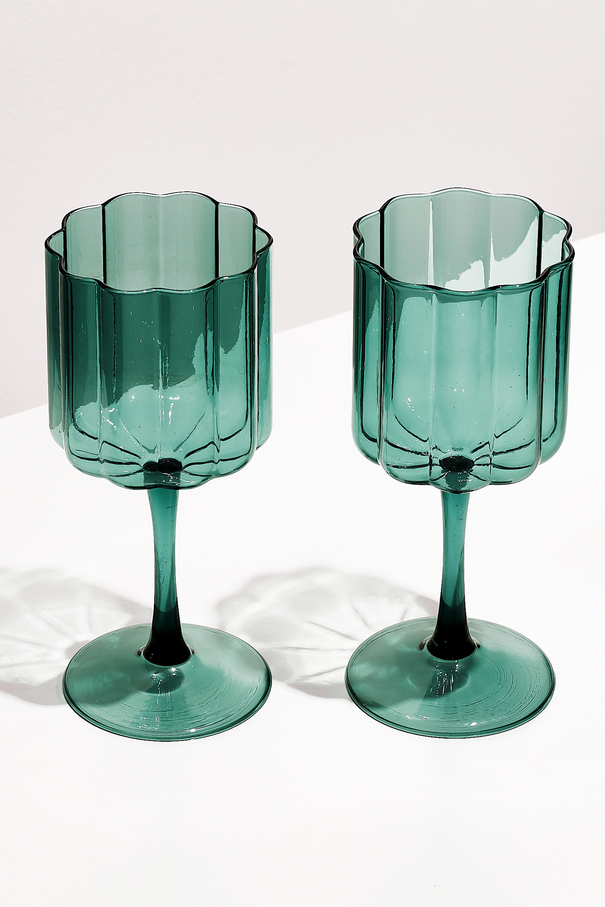 Two FAZEEK Teal Wave Wine Glasses with green-tinted, scalloped edges and textured sides, crafted from hand-blown borosilicate glass, rest on a white surface.