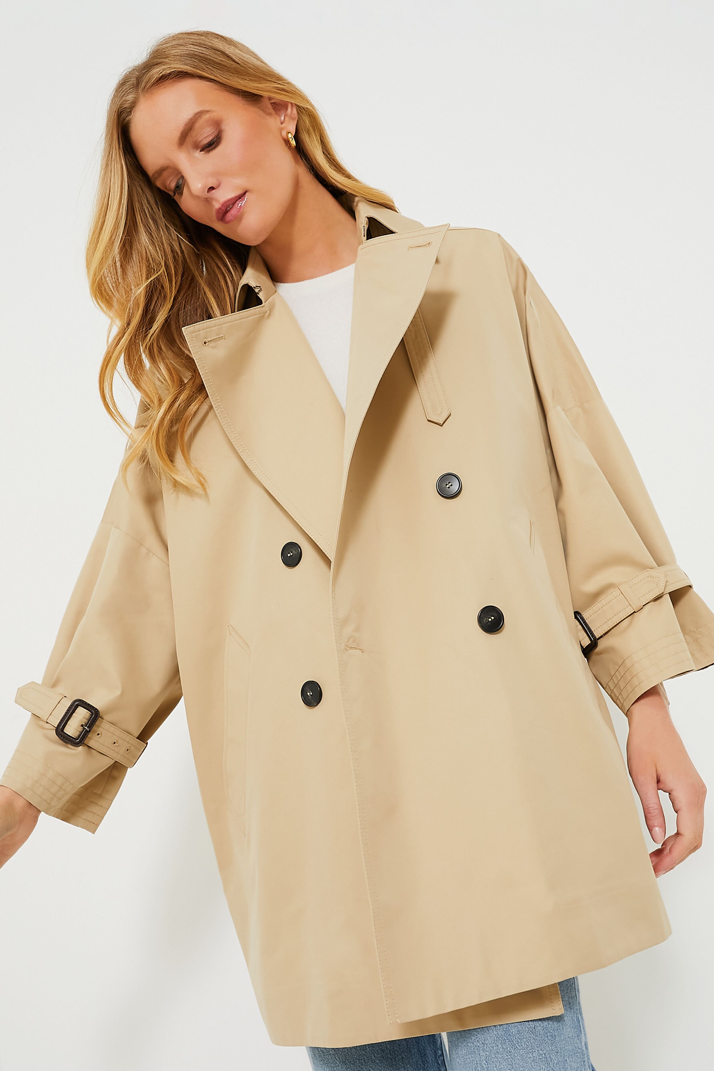 A woman wears a loose-fitting beige water-repellent trench coat with black buttons and cuff straps from Weekend Max Mara, paired with light blue jeans and a white top, embodying Italian elegance.