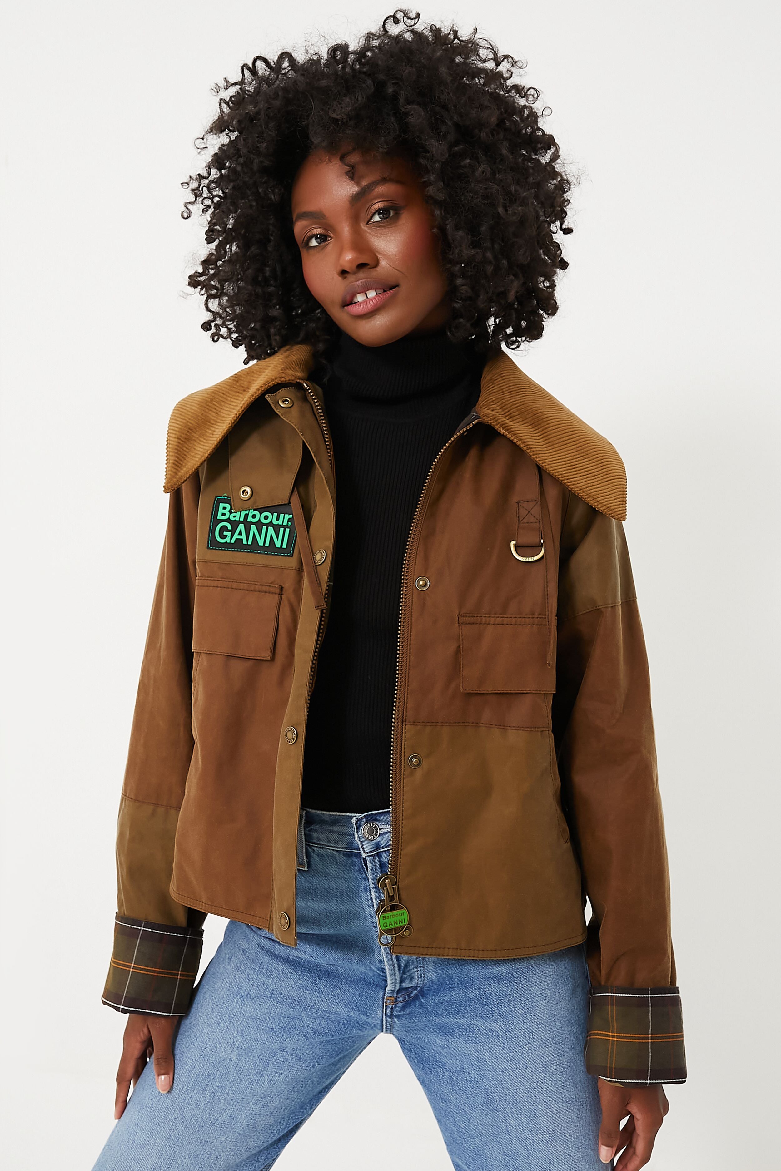 A woman stands confidently in a Barbour x GANNI jacket with an oversized collar, paired with a black turtleneck and blue jeans.
