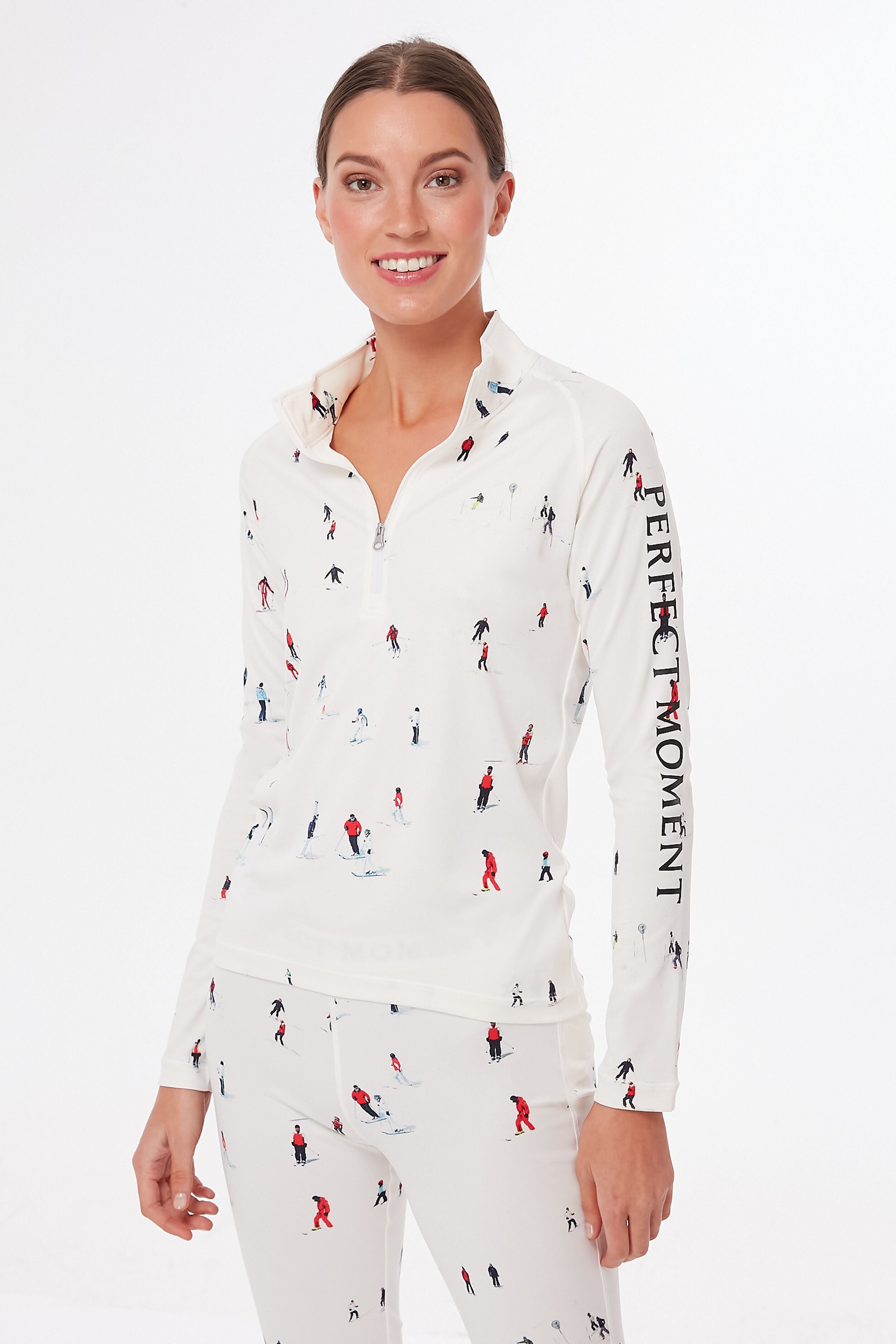 A woman wears a matching white Perfect Moment Ski Print Thermal Half Zip top and pants with small skier prints and PERFECT MOMENT text on the sleeve, ideal for après ski or chic base layering.