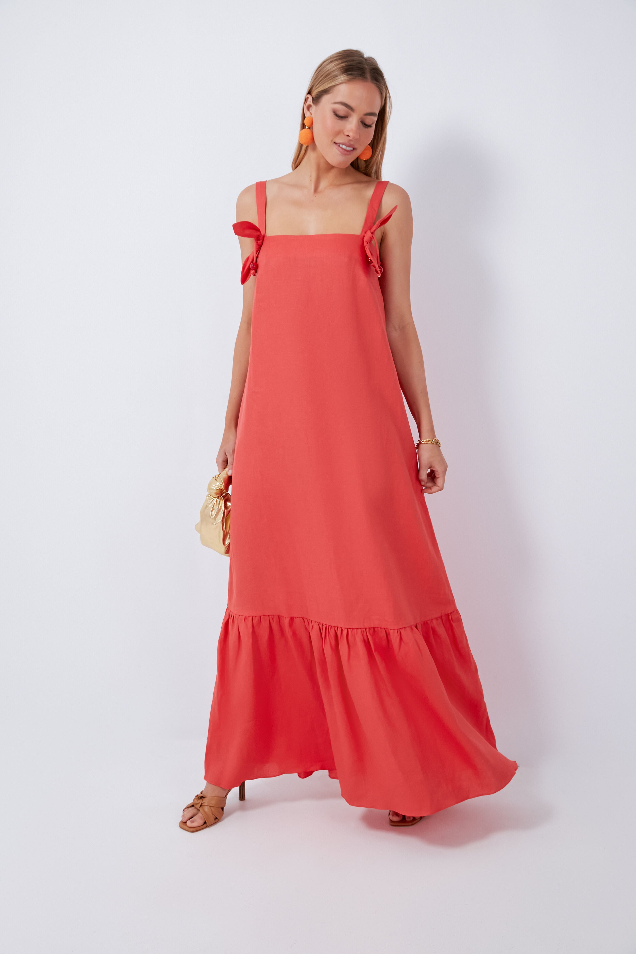 Woman in a coral sleeveless linen maxi dress with ruffle details by ADRIANA DEGREAS, styled with orange earrings, a bracelet, and sandals.