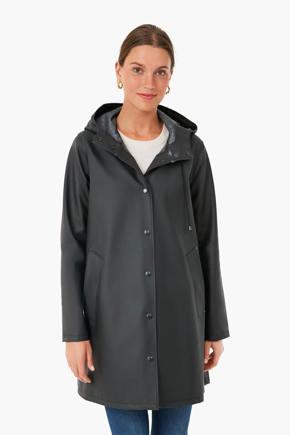 A woman faces forward, wearing a long black Stutterheim raincoat with a hood, paired with a white top and blue jeans.