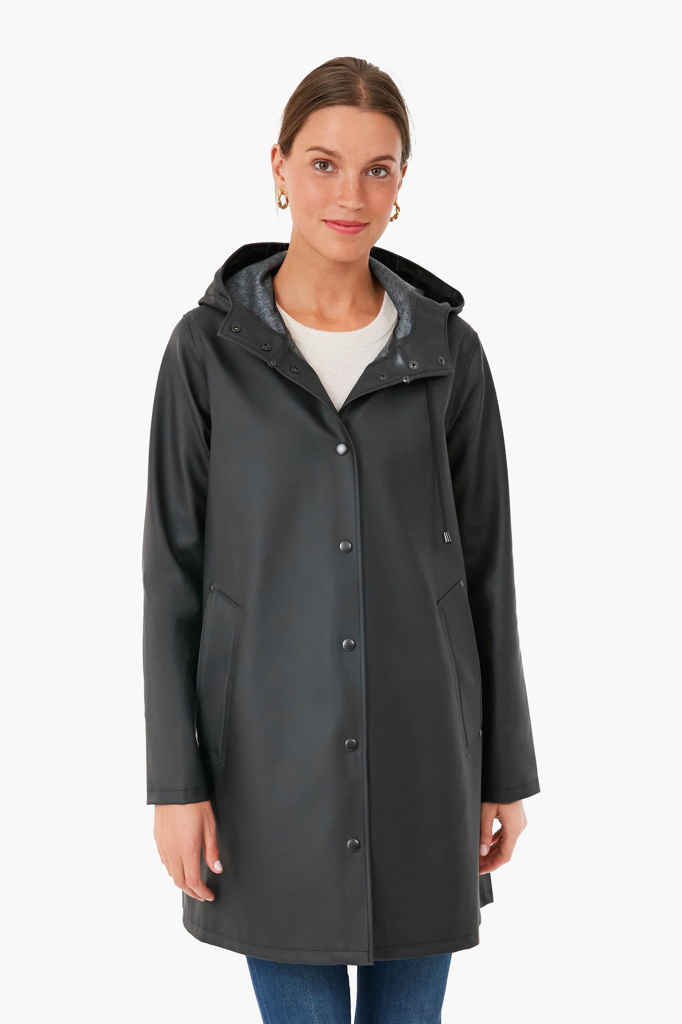 A woman faces forward, wearing a long black Stutterheim raincoat with a hood, paired with a white top and blue jeans.