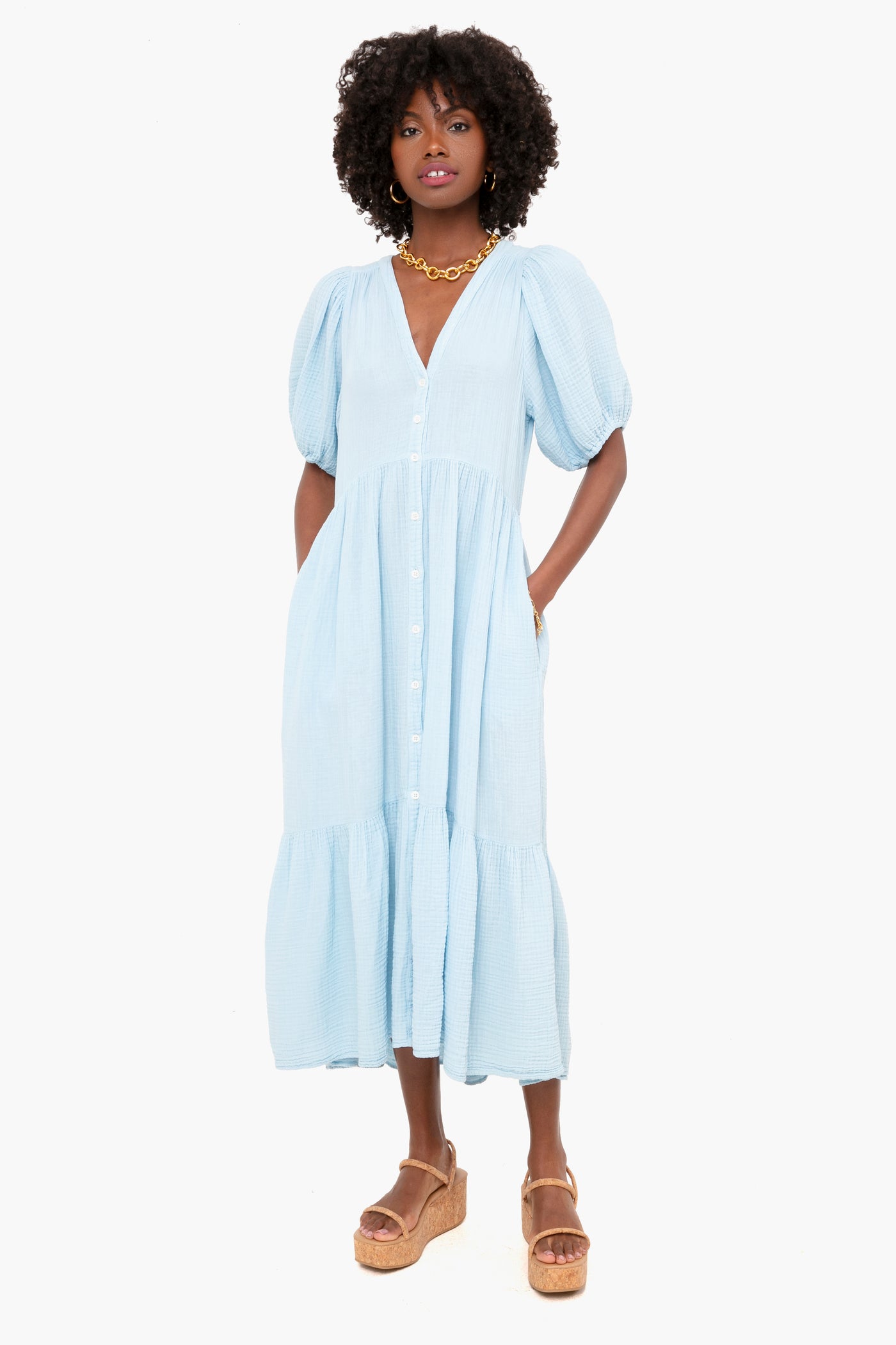 A woman wears the XiRENA Cyprus Blue Lennox Dress, a light blue cotton gauze midi with puff sleeves and a tiered skirt, styled with a gold necklace and tan platform sandals.