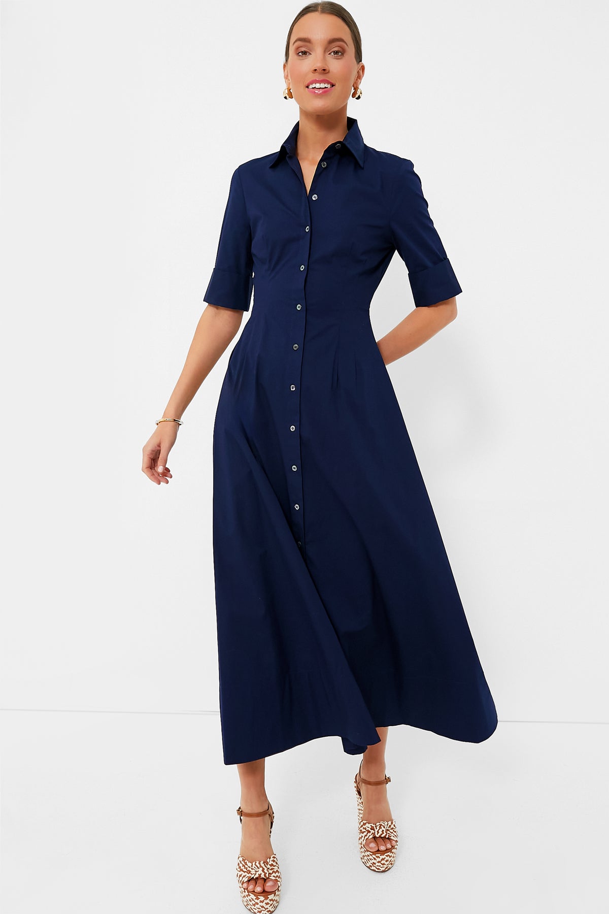 A woman wears the STAUD Joan Dress, a navy blue button-front collared midi dress with short sleeves, paired with beige heeled sandals.