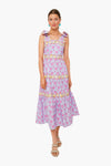 A woman wears the CeliaB Elara Dress—a sleeveless, light purple midi dress with pink and yellow floral patterns, ruffle shoulder details, and sandals—showcasing a vibrant, tropical-inspired look in sustainable fashion.