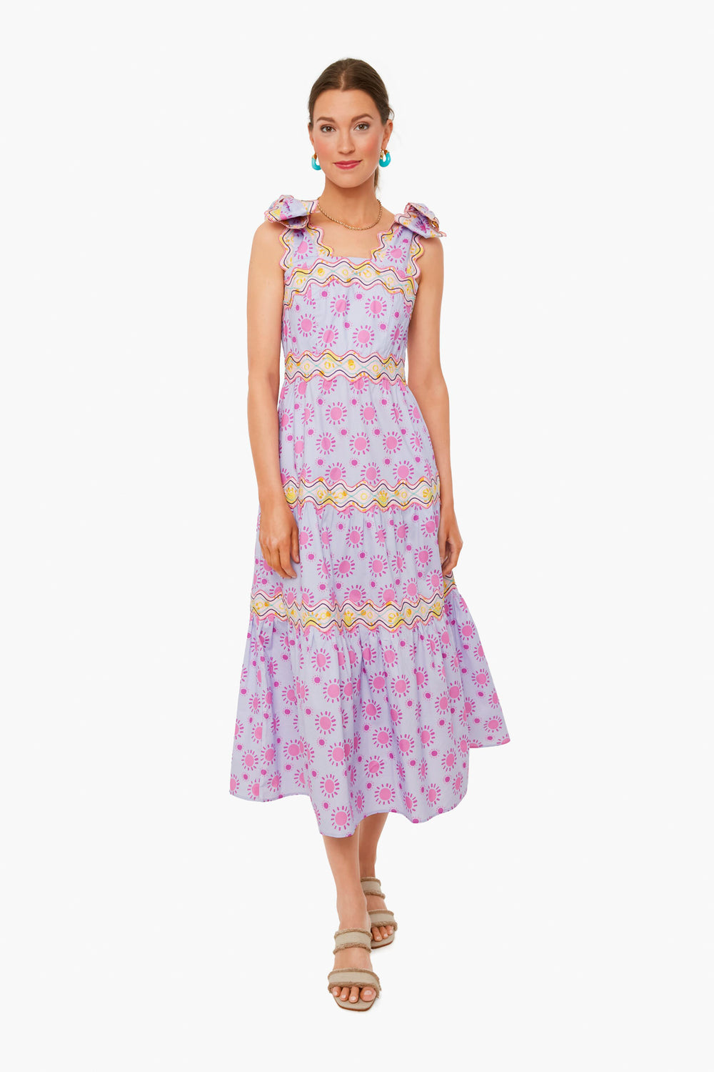 A woman wears the CeliaB Elara Dress—a sleeveless, light purple midi dress with pink and yellow floral patterns, ruffle shoulder details, and sandals—showcasing a vibrant, tropical-inspired look in sustainable fashion.