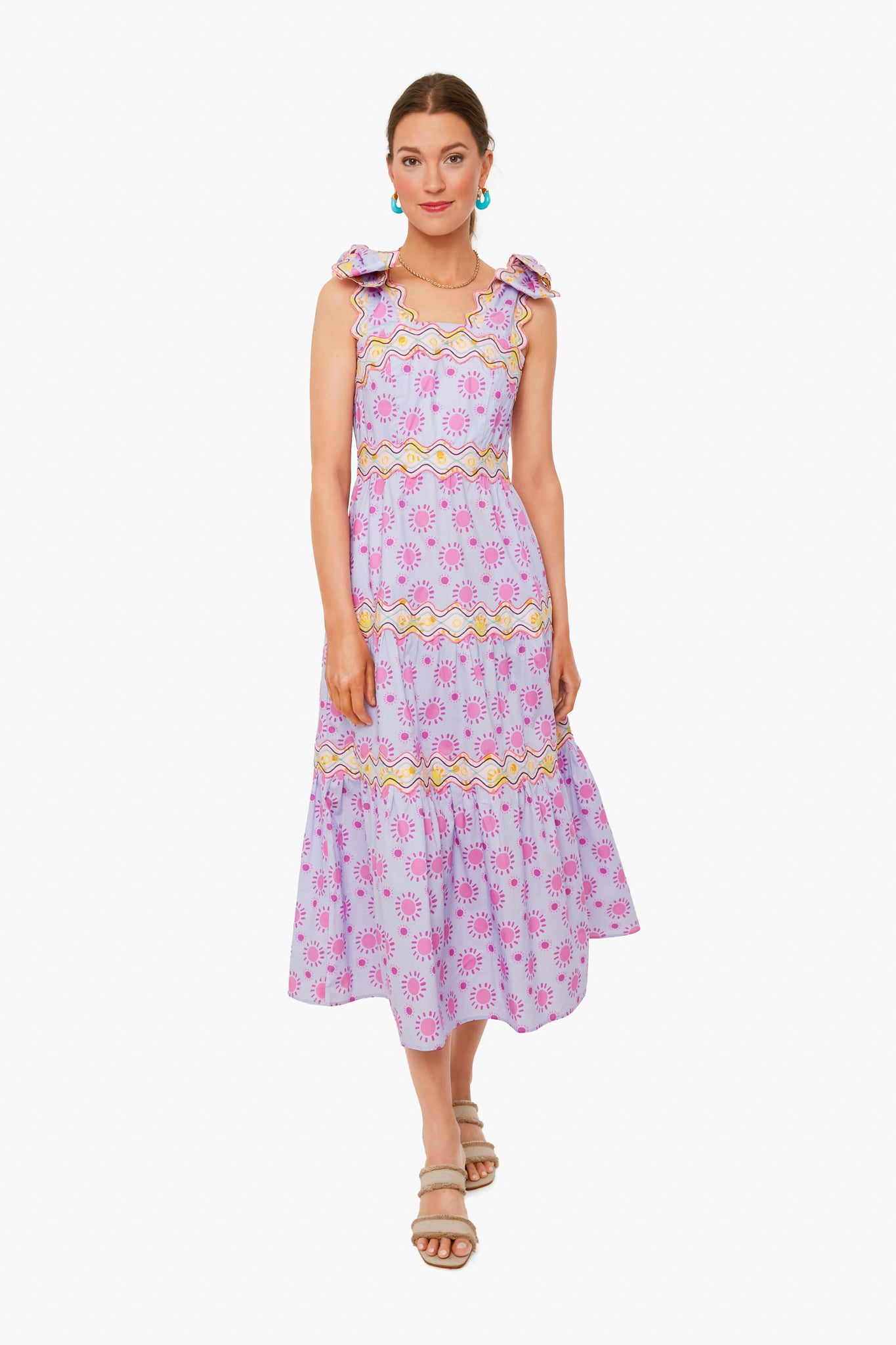 A woman wears the CeliaB Elara Dress—a sleeveless, light purple midi dress with pink and yellow floral patterns, ruffle shoulder details, and sandals—showcasing a vibrant, tropical-inspired look in sustainable fashion.