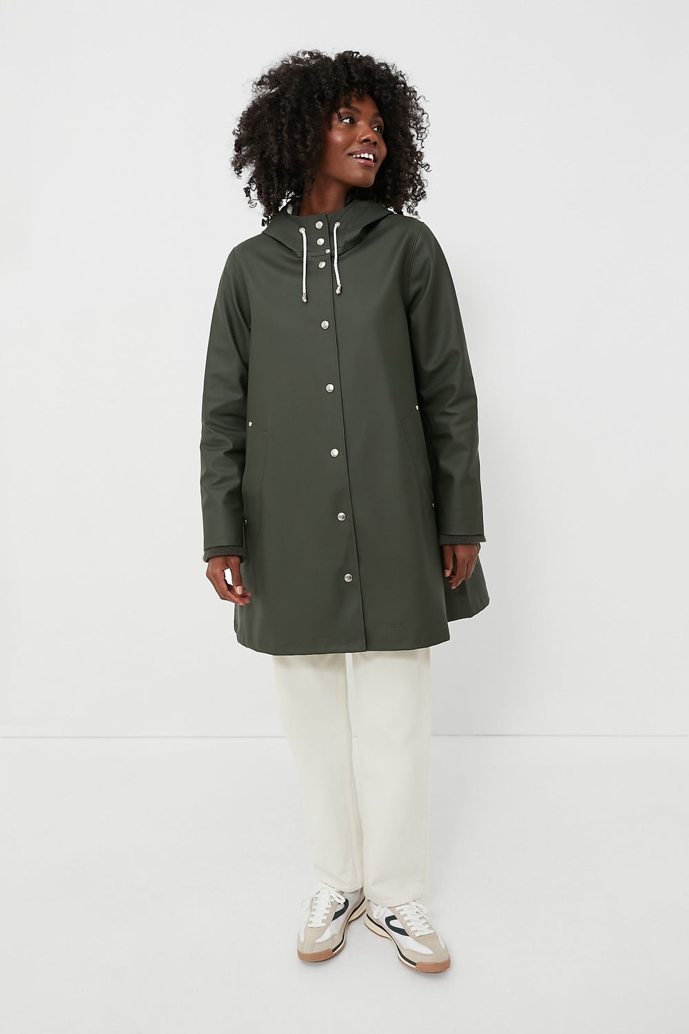 A person stands confidently in a dark green Stutterheim hooded A-line raincoat, paired with white pants and white sneakers.