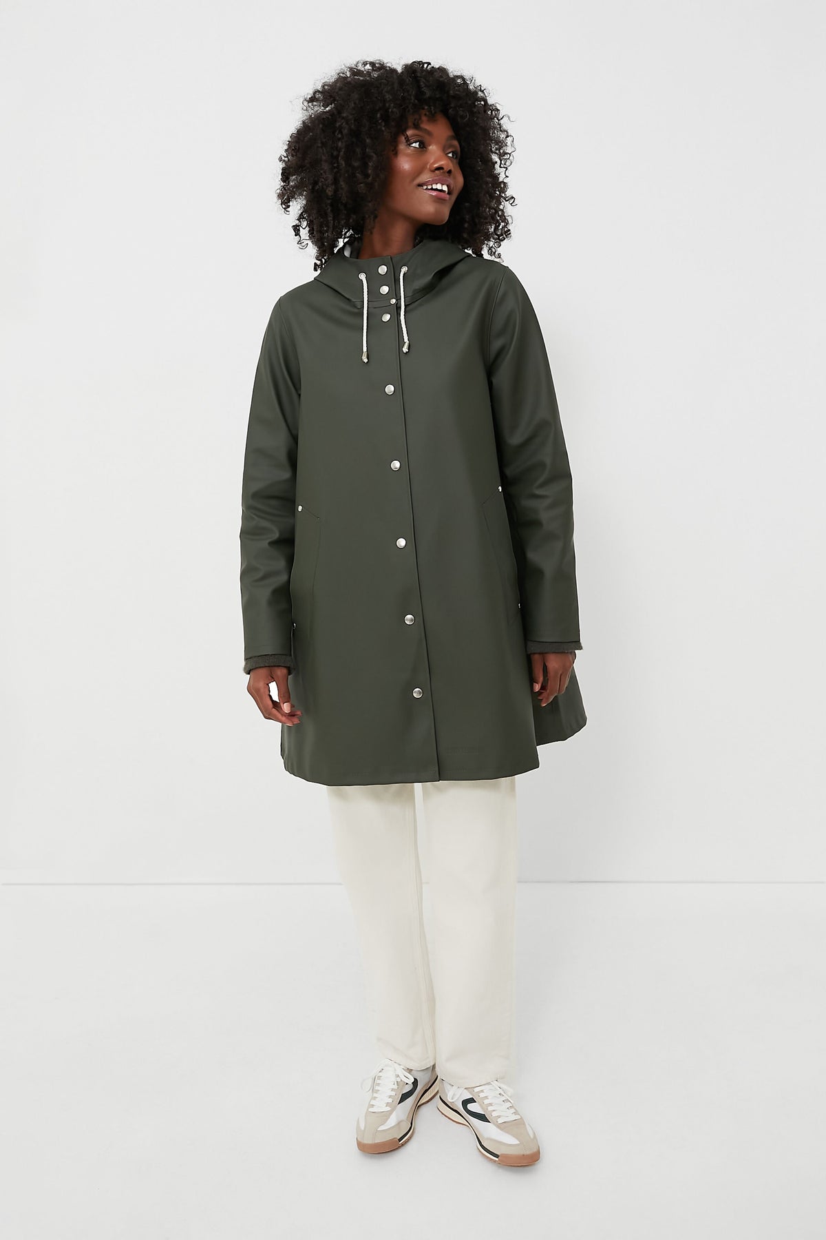 A person stands confidently in a dark green Stutterheim hooded A-line raincoat, paired with white pants and white sneakers.