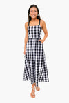 A woman stands smiling in a sleeveless black and white gingham dress with black straps from Tuckernuck, paired with hoop earrings and sandals—an ideal addition to any summer wardrobe.