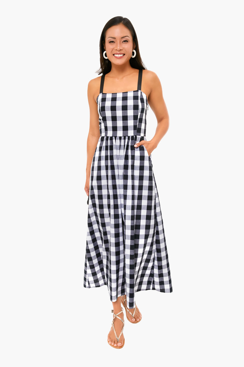 A woman stands smiling in a sleeveless black and white gingham dress with black straps from Tuckernuck, paired with hoop earrings and sandals—an ideal addition to any summer wardrobe.