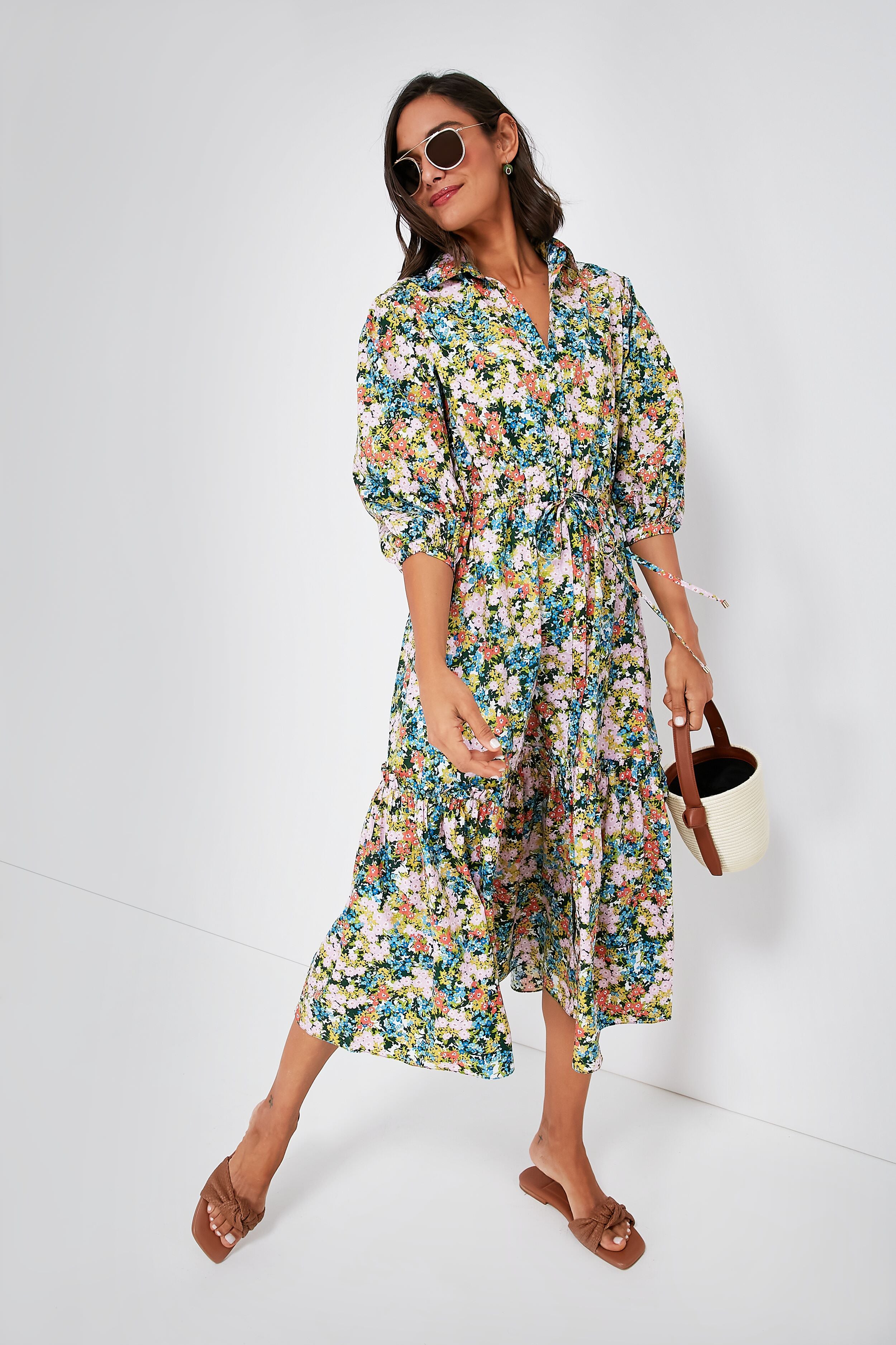 A woman in sunglasses wears a long romantic floral Cara Cara dress, brown sandals, and holds a small white and brown handbag, standing confidently.