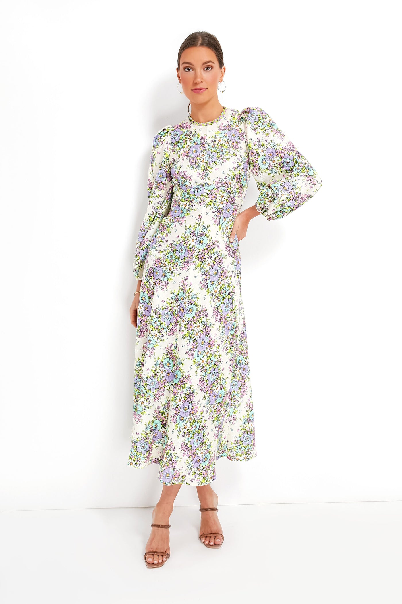 A woman wears an Alemais Petal Agnes Midi linen dress with a floral chevron print, puffed sleeves, and tan heeled sandals.