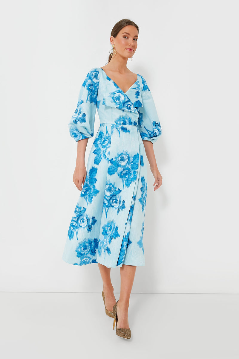 A woman wears the Emilia Wickstead Blue Gabby Dress, a light blue floral wrap dress with three-quarter puff sleeves, paired with beige heels.