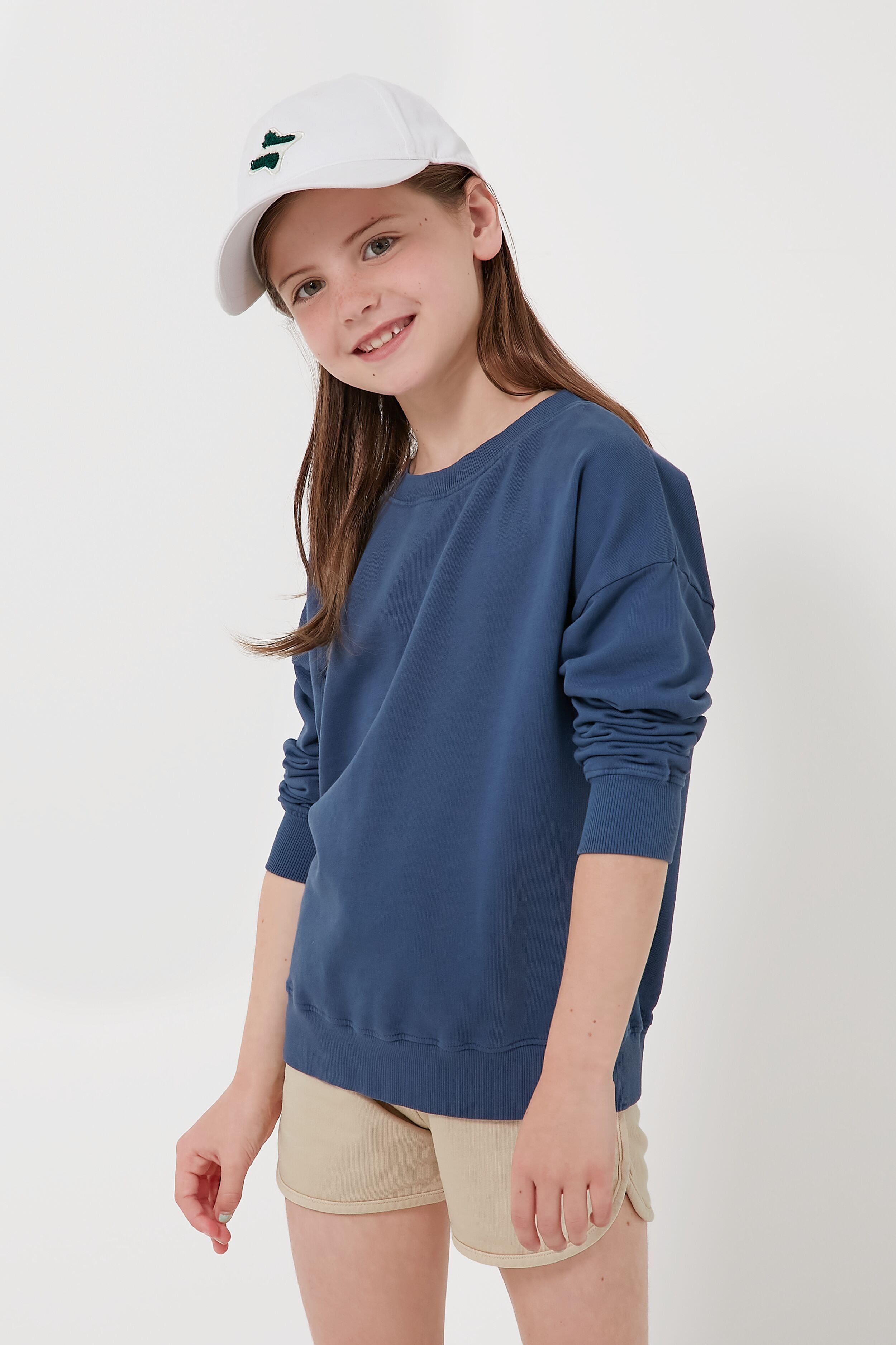 A girl in Les Gamins' organic cotton sweatshirt, beige shorts, and white cap smiles at the camera—stylish and sustainably made kids' clothing that’s comfortable too.