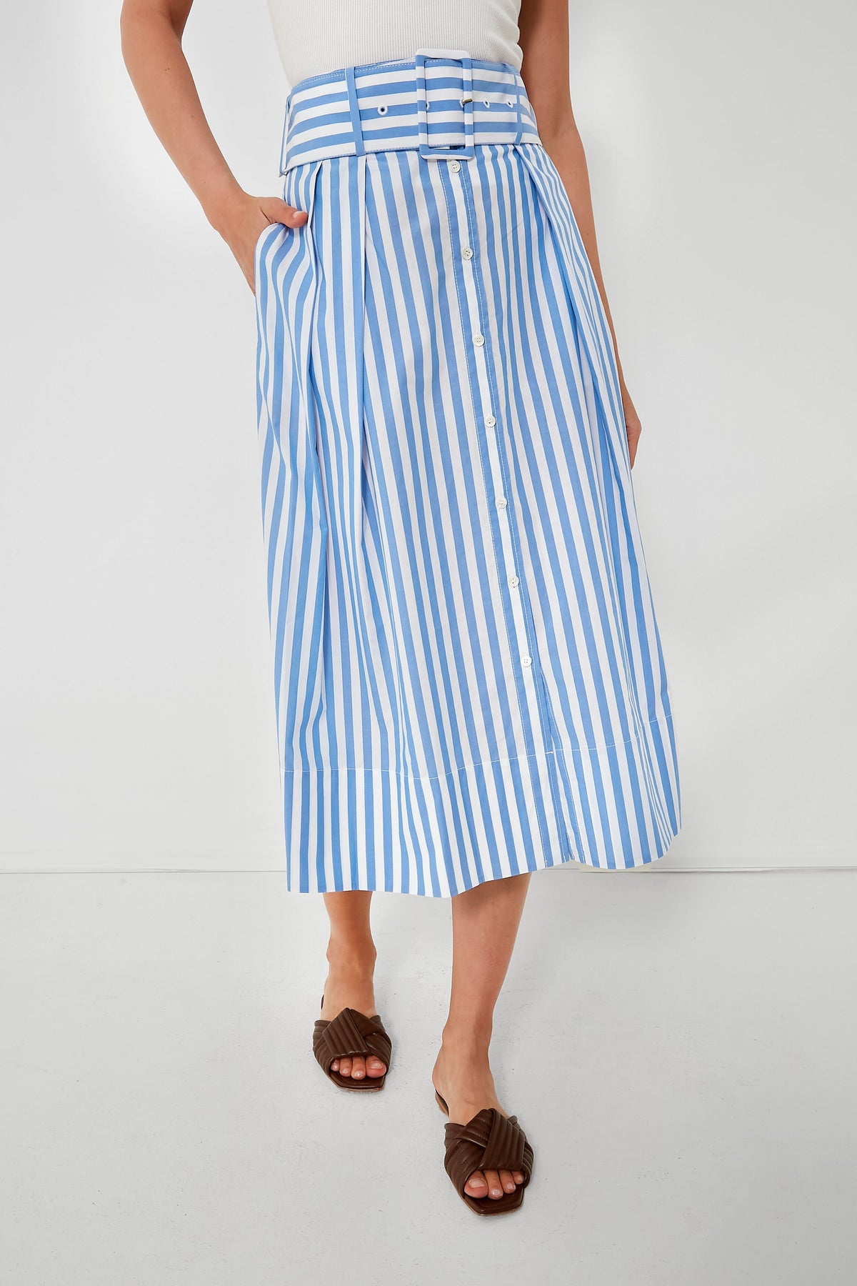 A person wears a STAUD cotton poplin high rise midi skirt with blue and white vertical stripes, front buttons, a matching belt, a white top, and brown sandals.