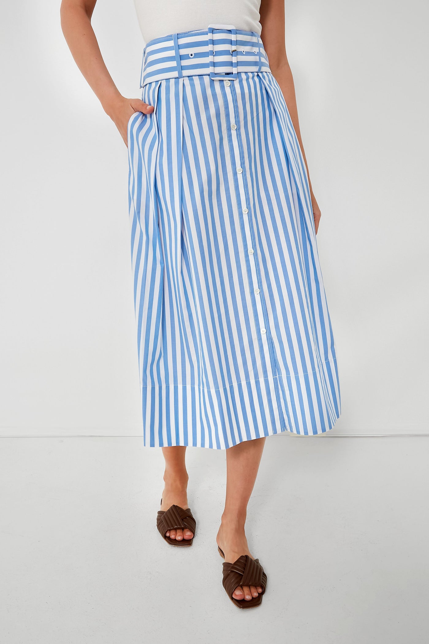 A person wears a STAUD cotton poplin high rise midi skirt with blue and white vertical stripes, front buttons, a matching belt, a white top, and brown sandals.