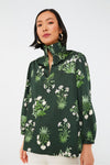 A woman wears a Tuckernuck dark green botanical print blouse with a high ruffle collar and three-quarter sleeves, paired with white pants.