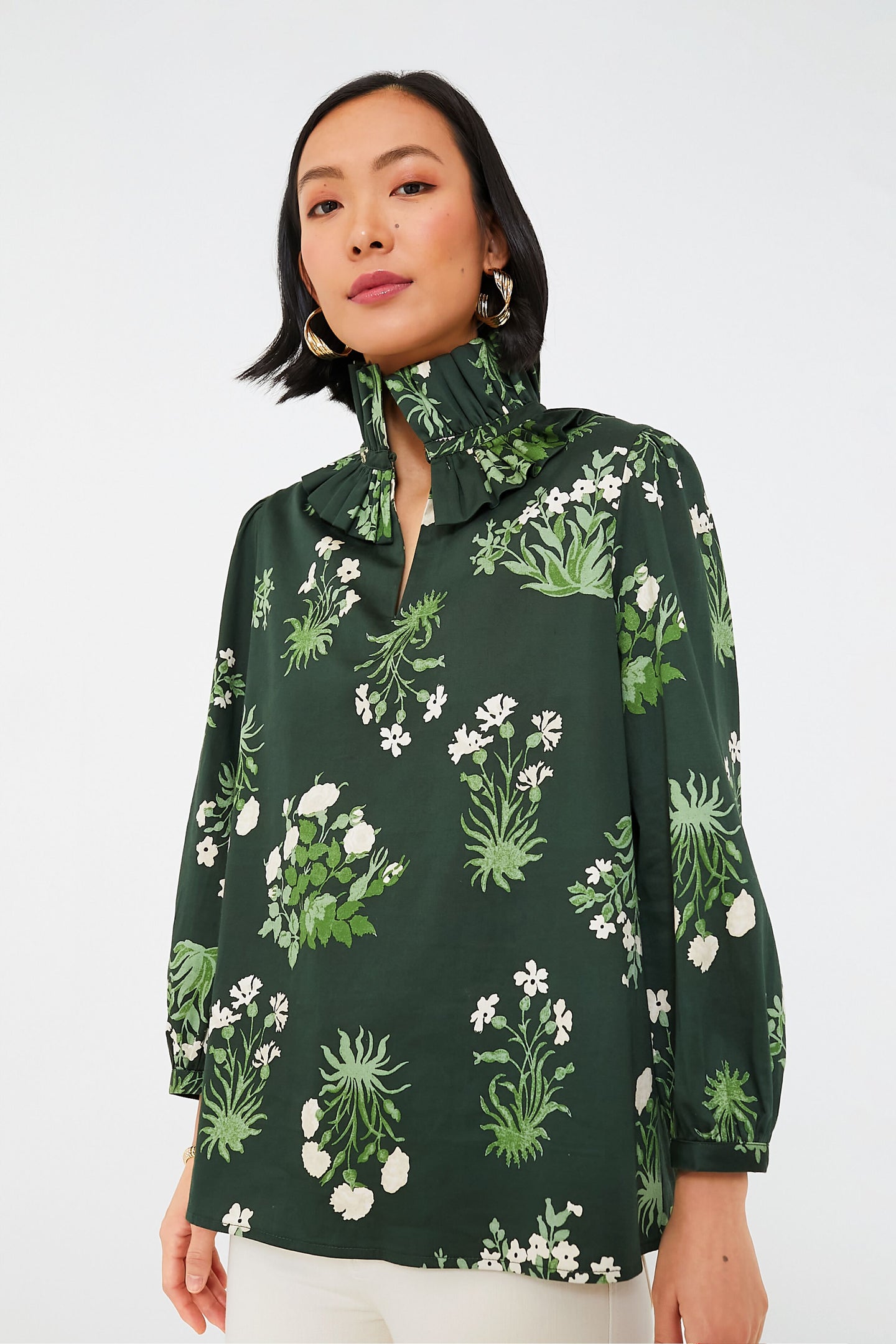 A woman wears a Tuckernuck dark green botanical print blouse with a high ruffle collar and three-quarter sleeves, paired with white pants.