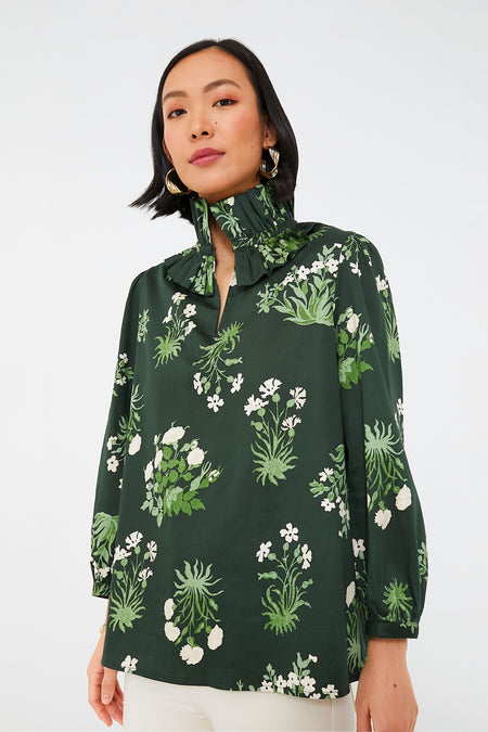 A woman wears a Tuckernuck dark green botanical print blouse with a high ruffle collar and three-quarter sleeves, paired with white pants.