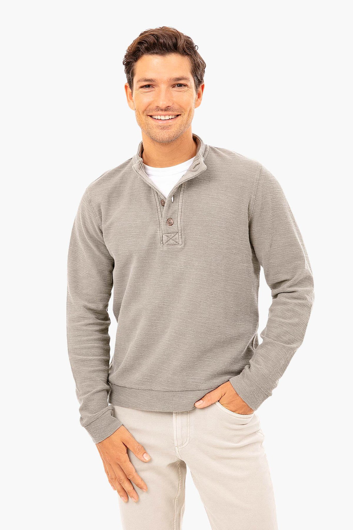 A man smiles, wearing a light gray Marine Layer eco-friendly sweater with a buttoned collar over a white shirt and light-colored pants.