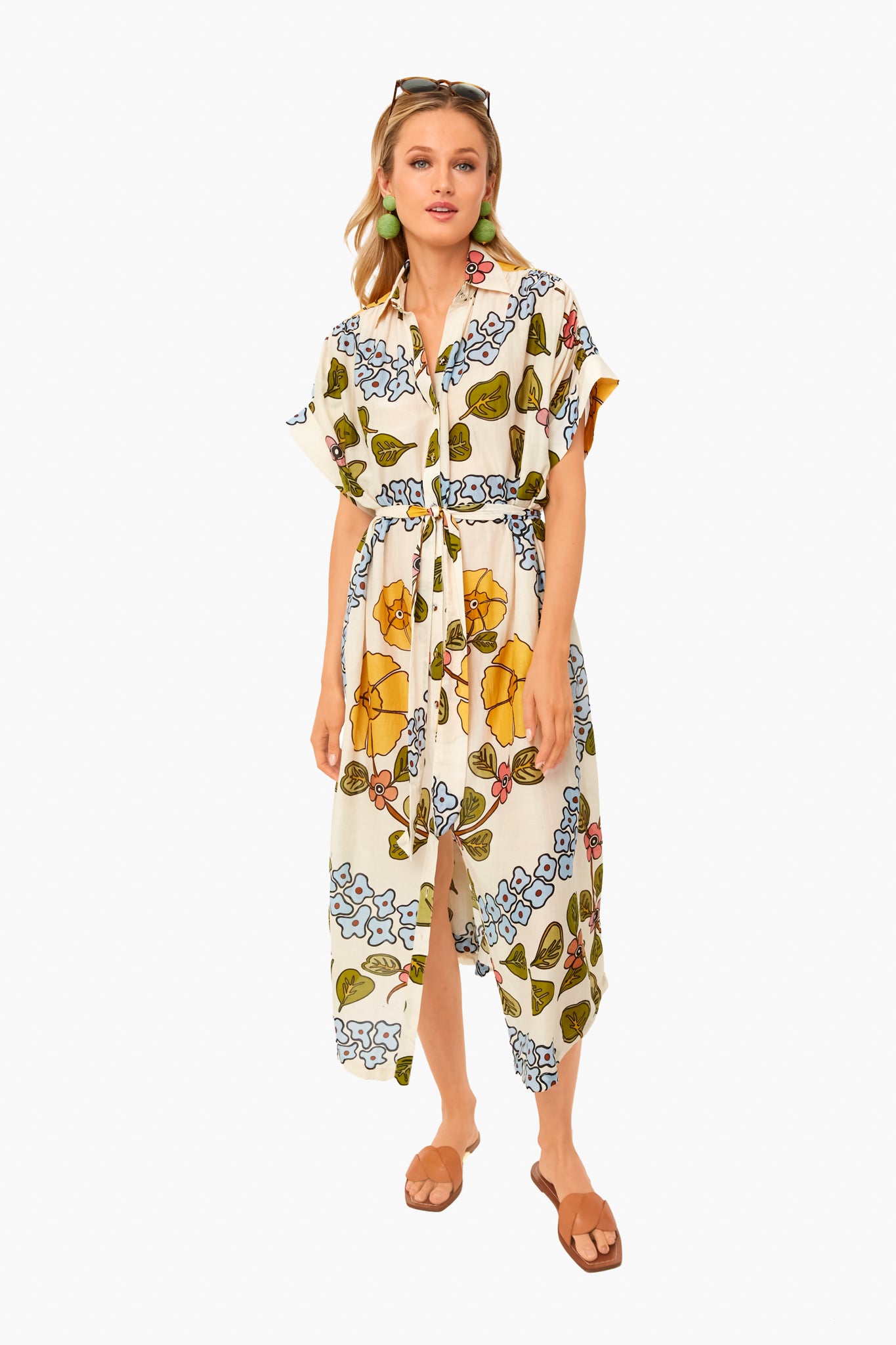 A woman models a Carolina K. belted, short-sleeve, button-down dress in a colorful floral cotton-silk blend, paired with green earrings, sunglasses on her head, and brown sandals.