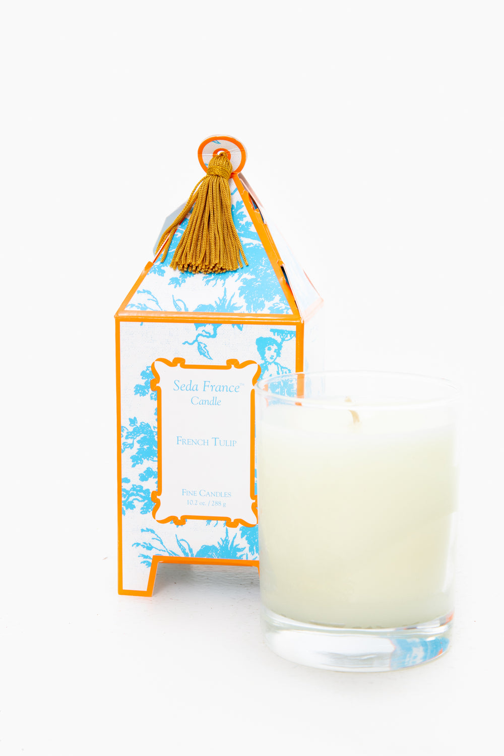 A Seda France glass jar candle sits before a blue and orange French Tulip Candle box adorned with a gold tassel, making it an ideal home fragrance or gift.