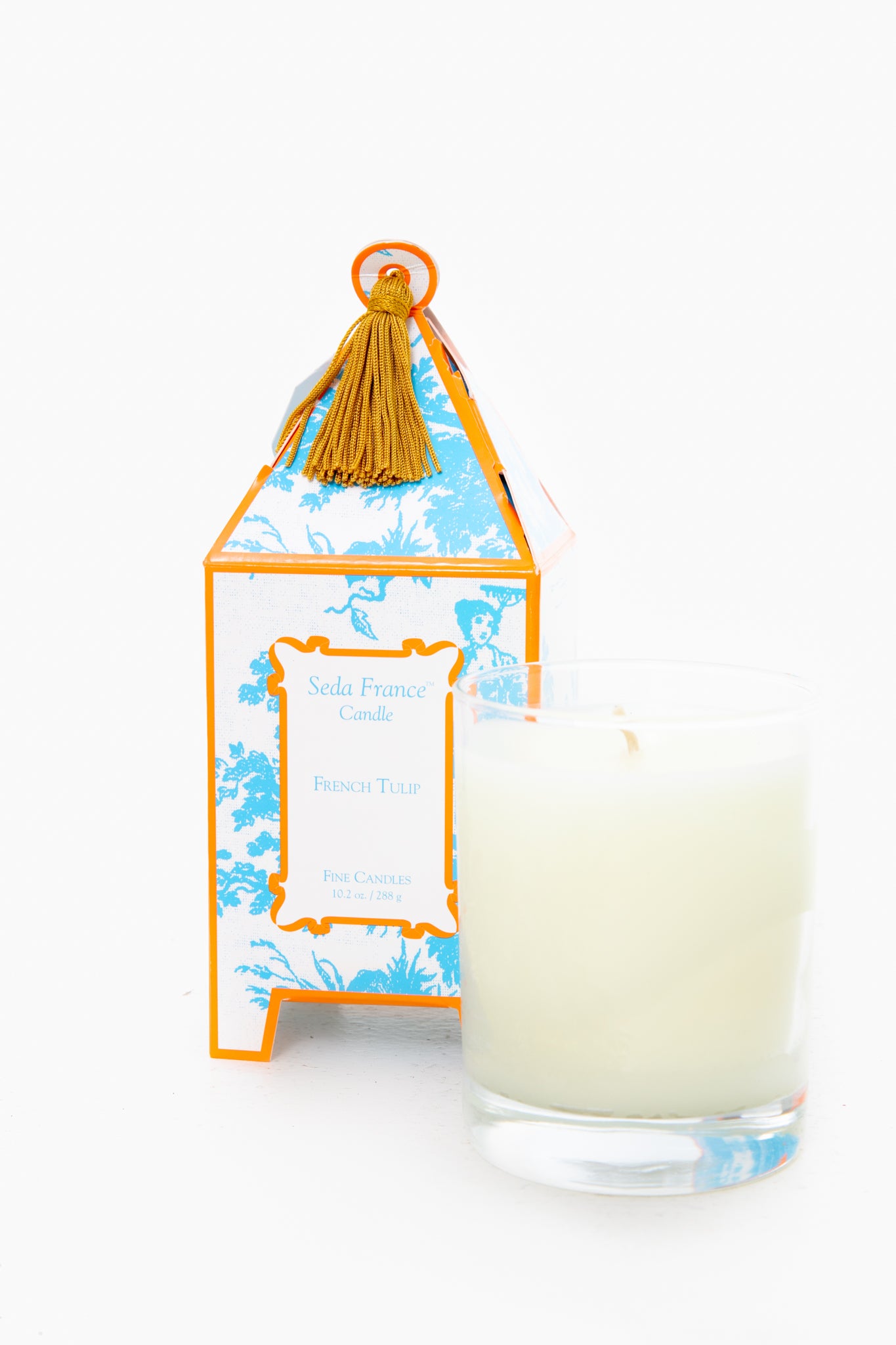 A Seda France glass jar candle sits before a blue and orange French Tulip Candle box adorned with a gold tassel, making it an ideal home fragrance or gift.