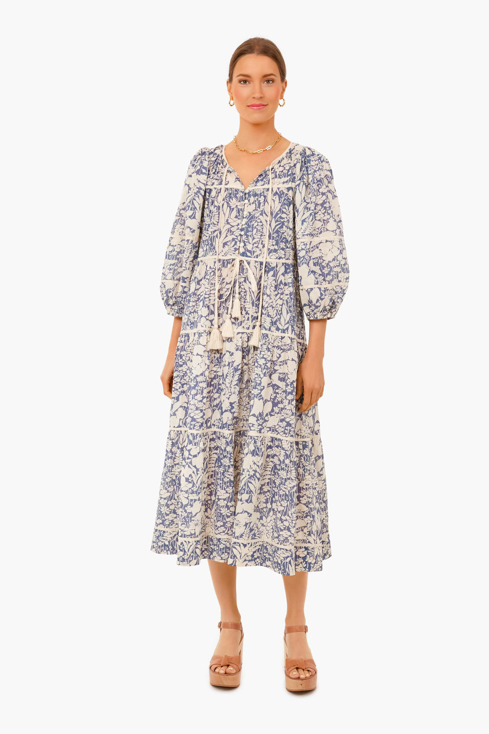 A woman faces forward in THE GREAT. Tassel Tie Blouson Dress with blue and white patterns and puff sleeves, paired with tan platform sandals.
