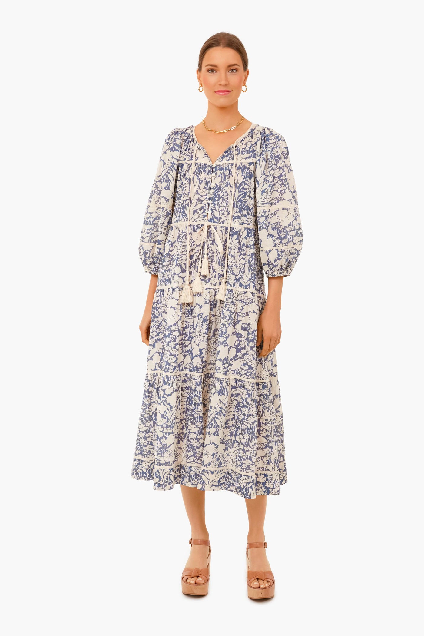 A woman faces forward in THE GREAT. Tassel Tie Blouson Dress with blue and white patterns and puff sleeves, paired with tan platform sandals.
